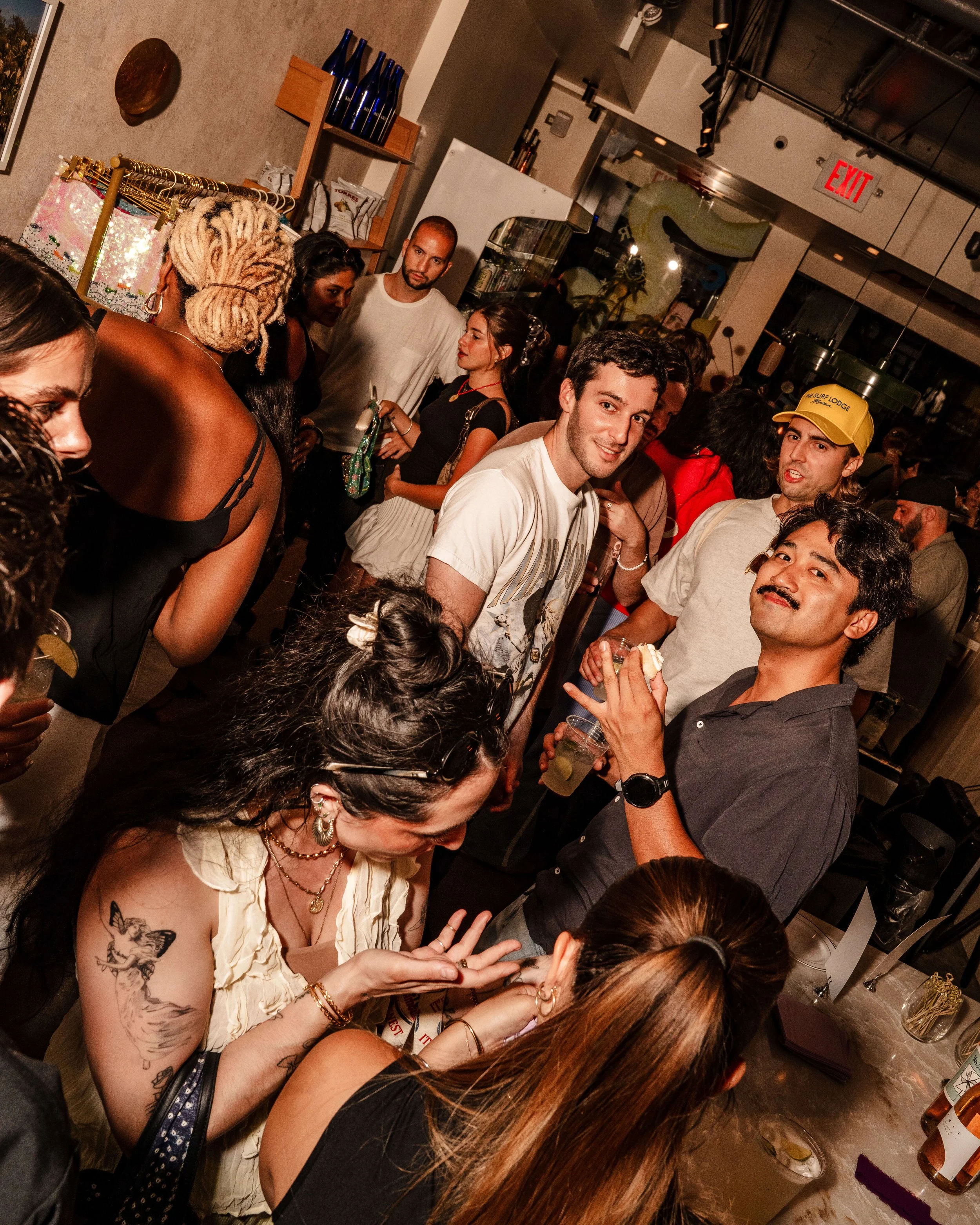 A group of young people gathered at a social event in a bar or restaurant, engaging in conversations and enjoying drinks. The scene is lively with a mix of men and women, some smiling and talking, with a busy background including shelves, bottles, an