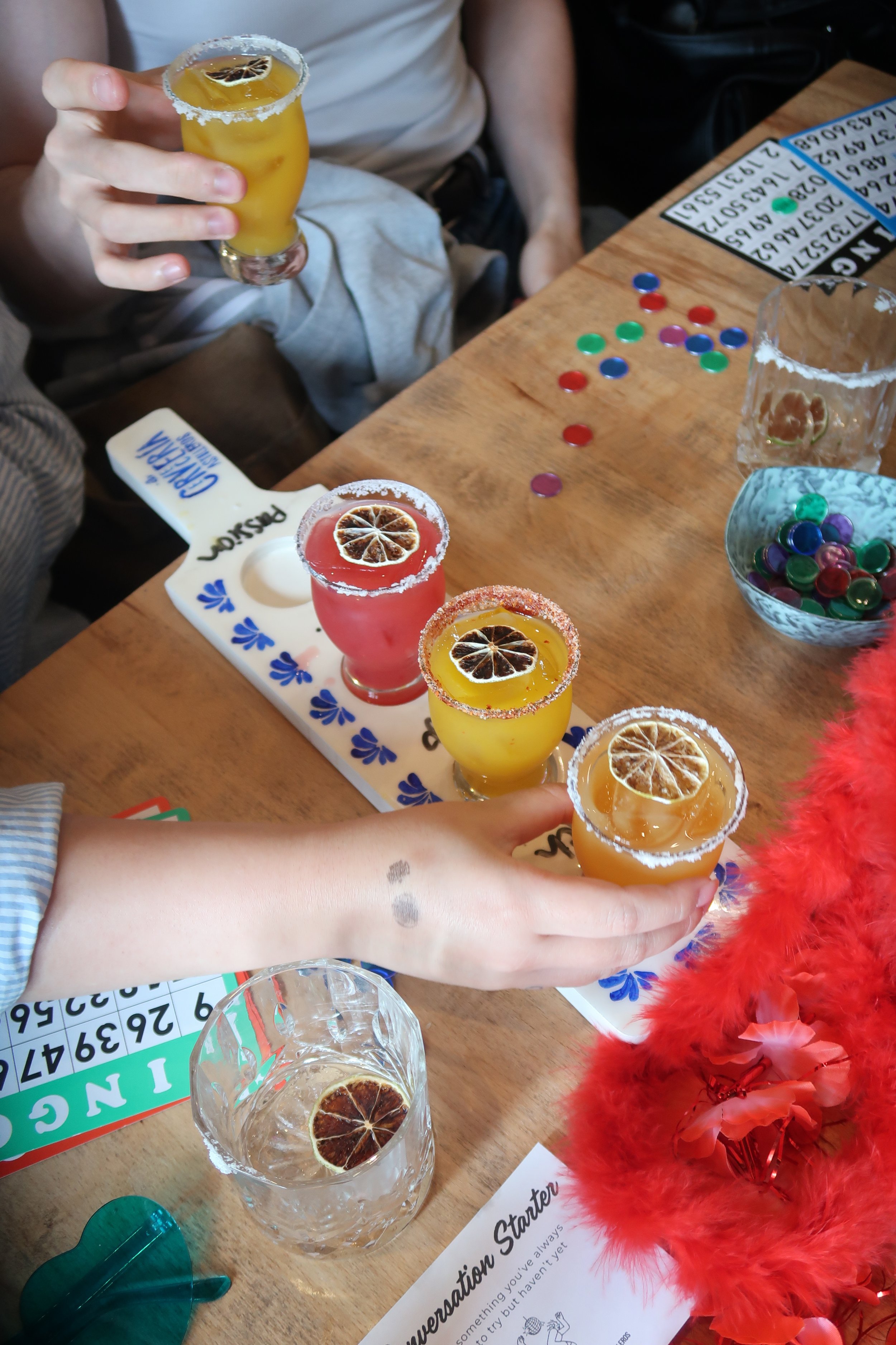 A group of people celebrating with four colorful cocktails topped with dried citrus slices, salt-rimmed glasses, a Bingo game, colorful tokens, and festive decorations on a wooden table.