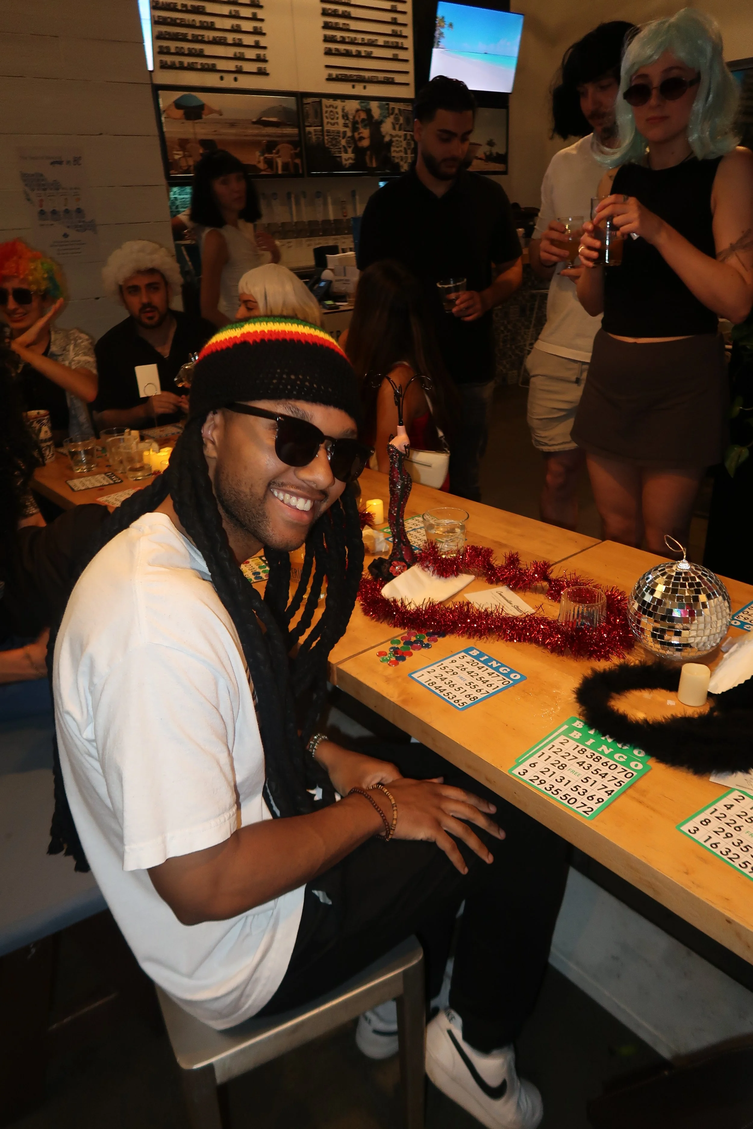 People celebrating a party with a man sitting at a table decorated with tinsel, a disco ball, and bingo cards, while others stand and socialize in the background. Some individuals wear colorful wigs and costumes.