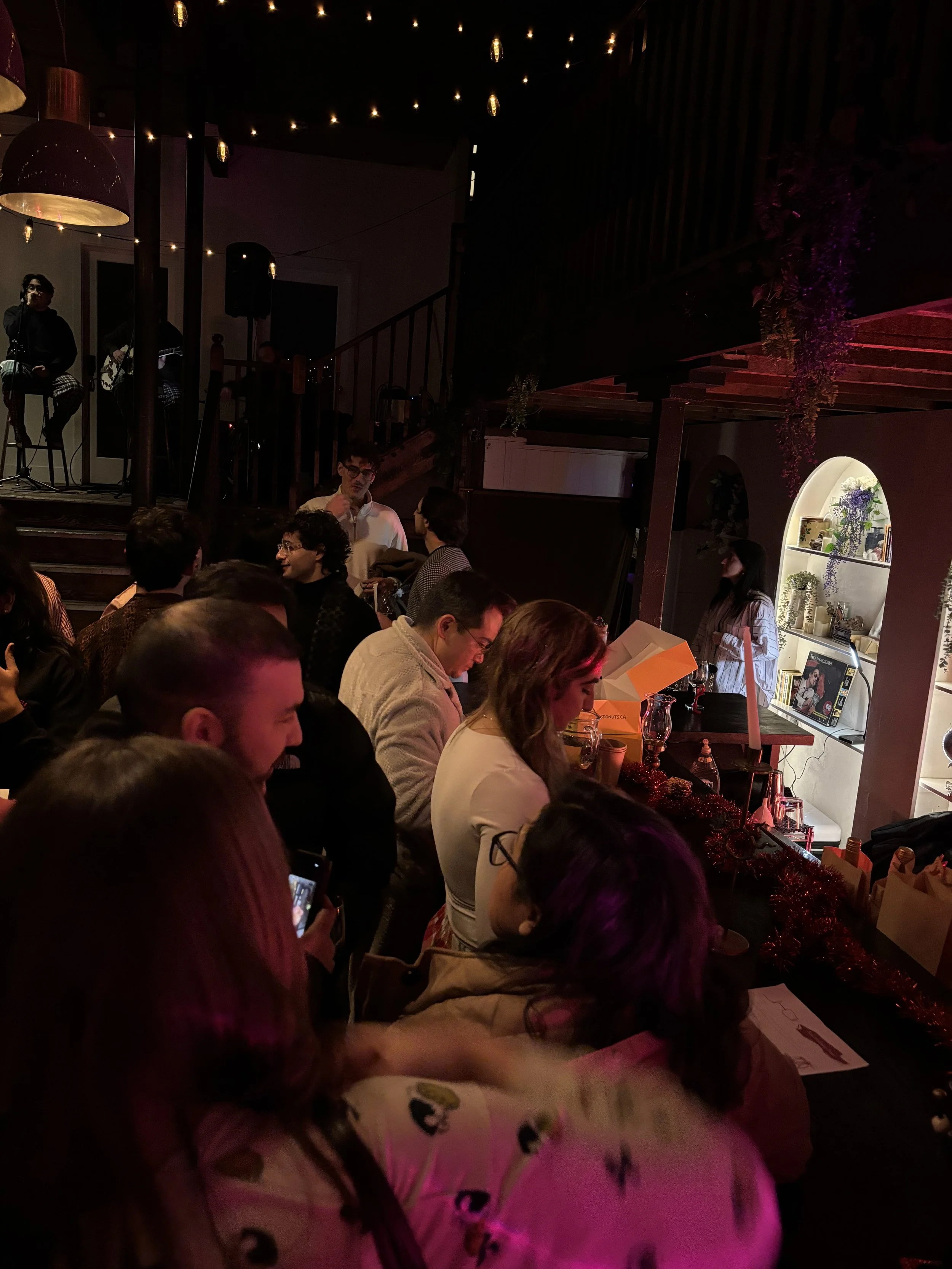People sitting at a bar or stage area, watching and listening to musicians on a small stage in a cozy, dimly lit venue with string lights overhead.