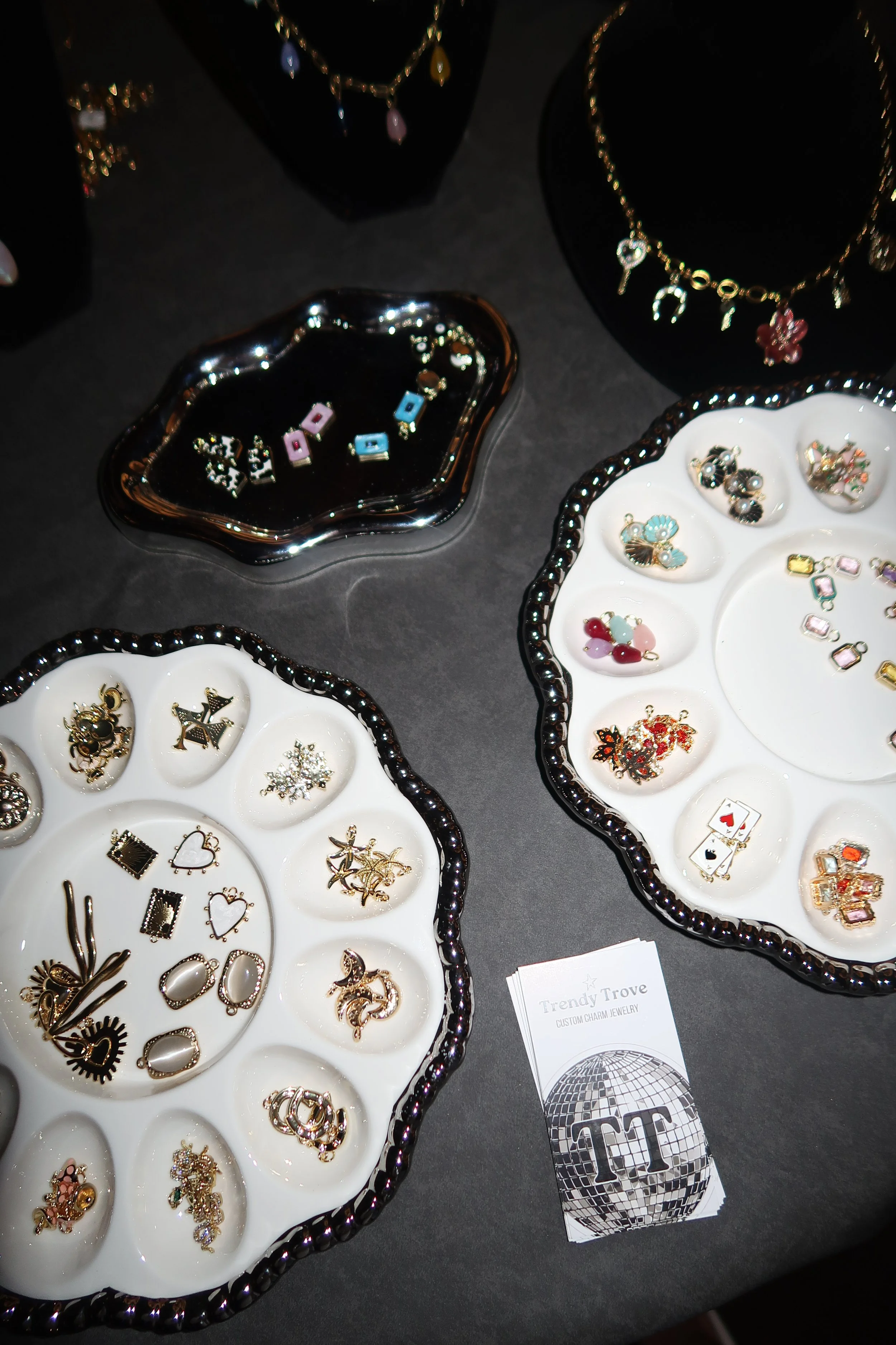 Jewelry display featuring charm earrings and necklaces on black and white trays, with business cards labeled 'Trendy Trove Custom Charm Jewelry'.