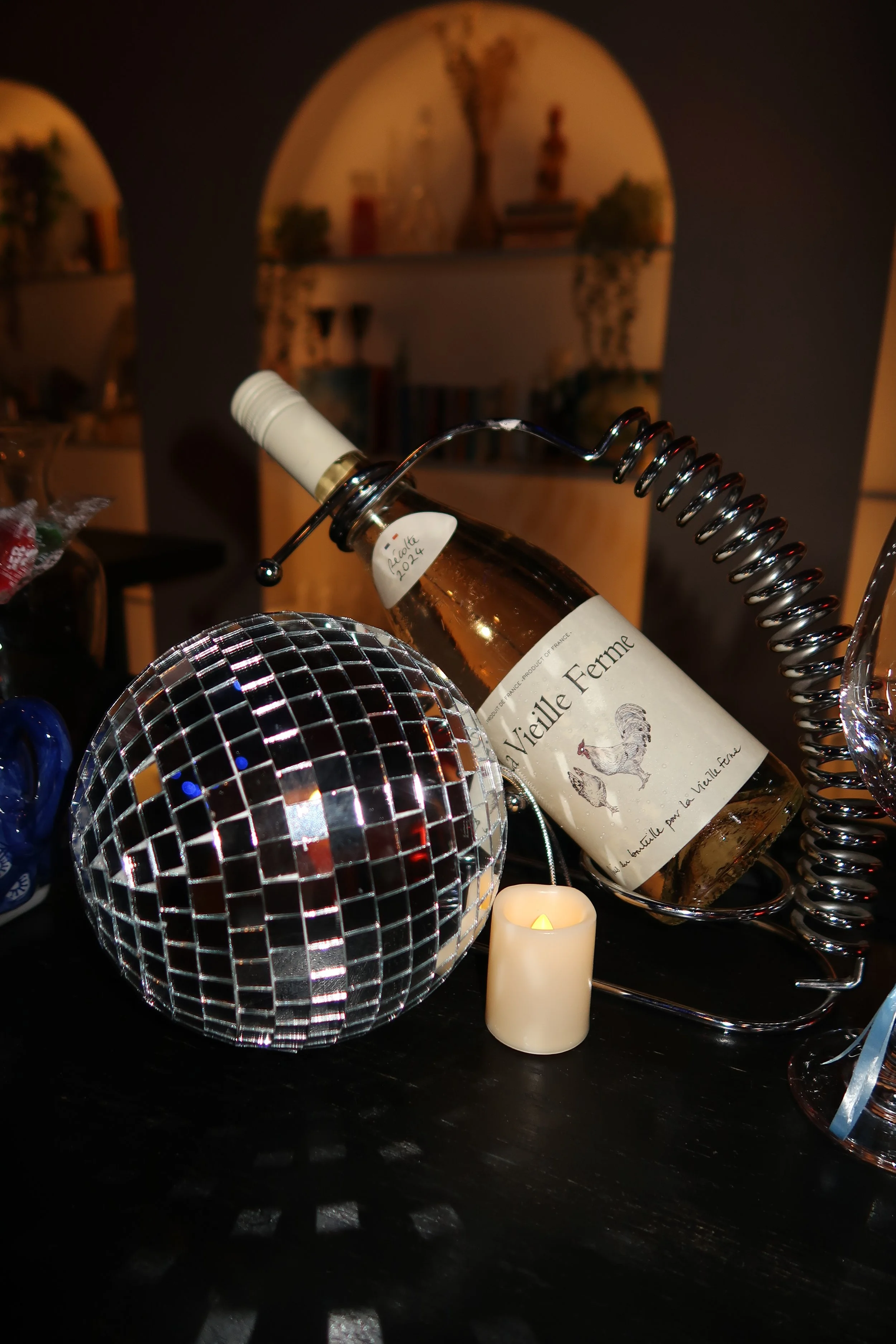Decorative scene including a leaning bottle of wine labeled 'La Vieille Ferme' in a metal wine holder, a small faux candle, a mirrored disco ball, and a glass on a dark surface in a dimly lit room.