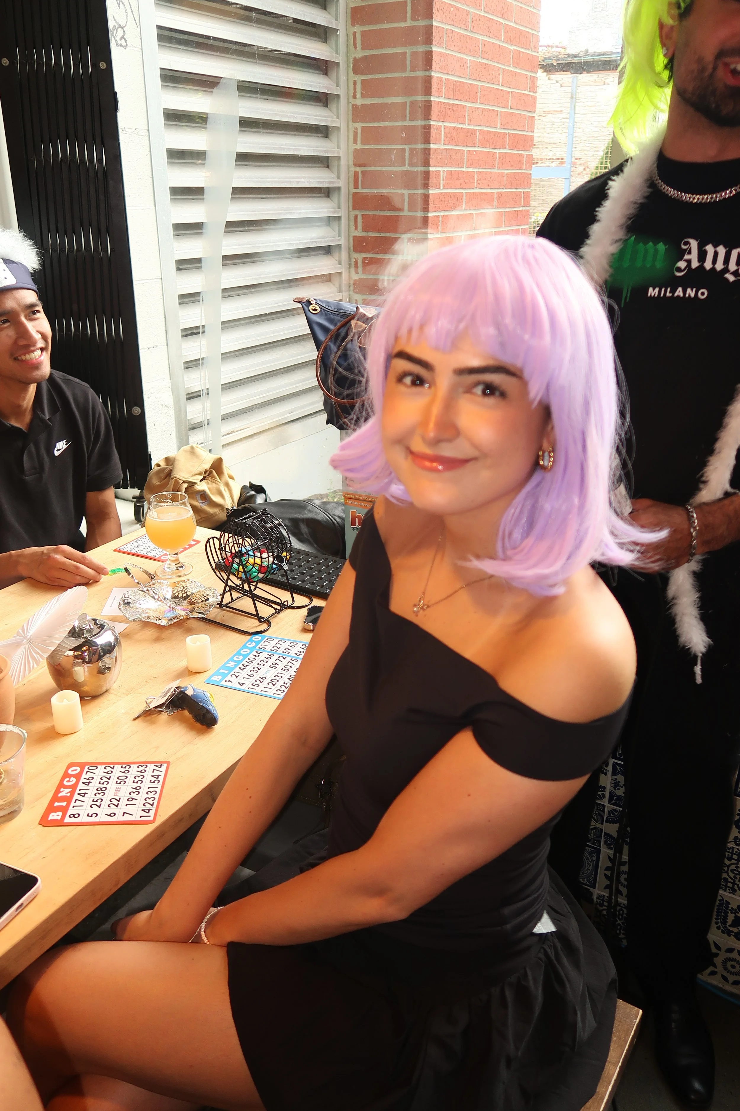 Young woman with pastel lavender hair smiling and sitting at a table with bingo cards, drinks, and game items, with two men nearby, one wearing a neon wig, in a room with brick and metal walls.