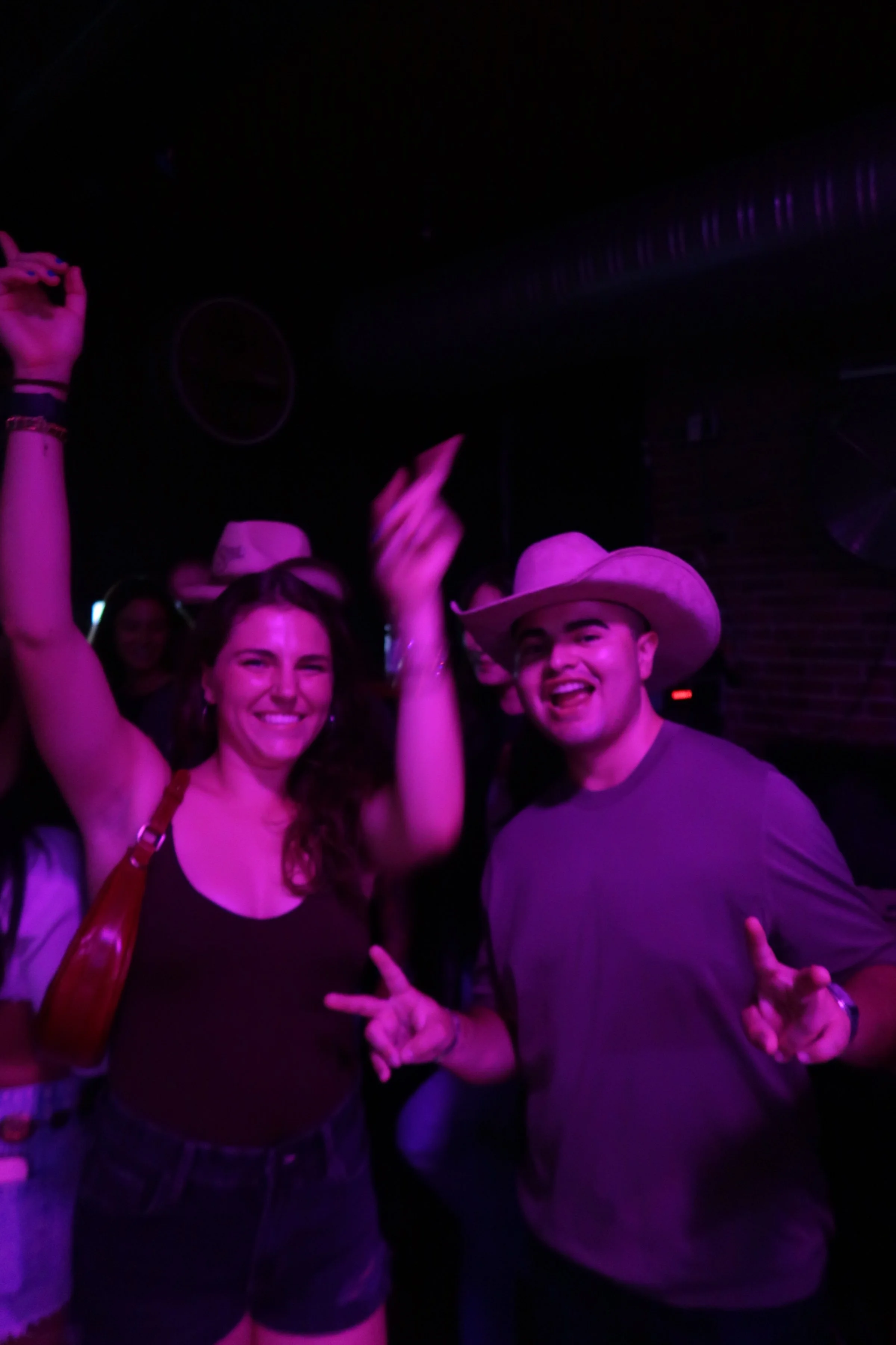 Two people smiling and posing for the camera in a dark, purple-lit setting, with one woman raising her arm and a man wearing a cowboy hat making peace signs.
