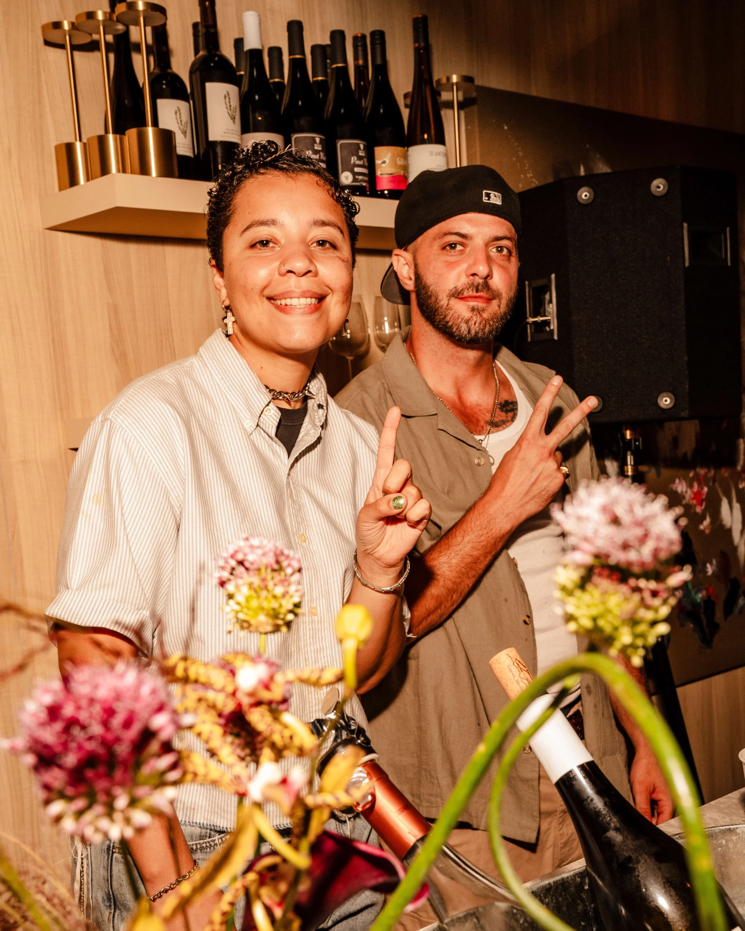 Two people smiling and making peace signs at a social gathering, with wine bottles on a shelf behind them and flowers in the foreground.