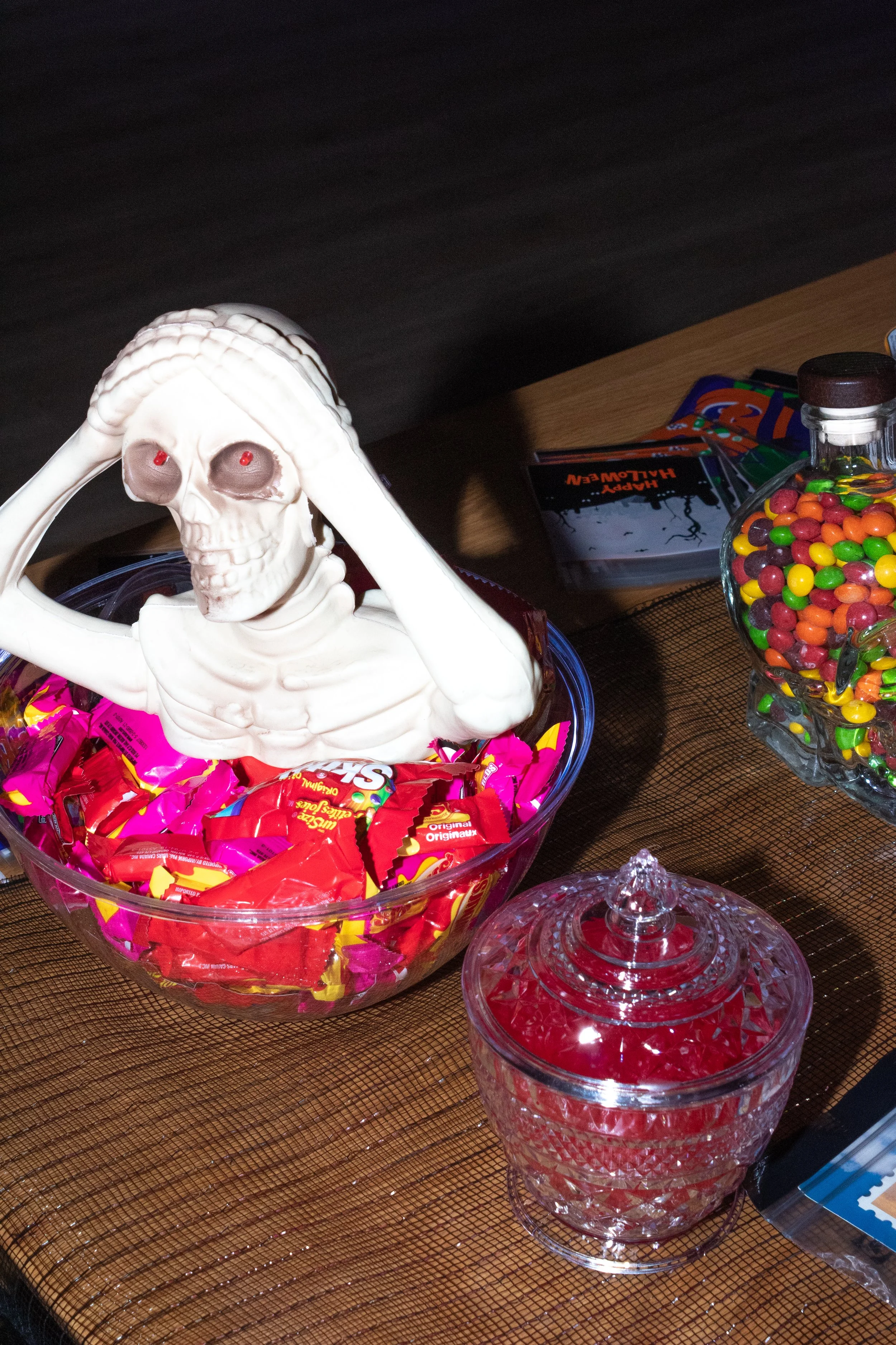 A skeleton-shaped Halloween decoration with red eyes, placed in a glass bowl filled with pink and red candies, on a table with other candies and Halloween-themed items.