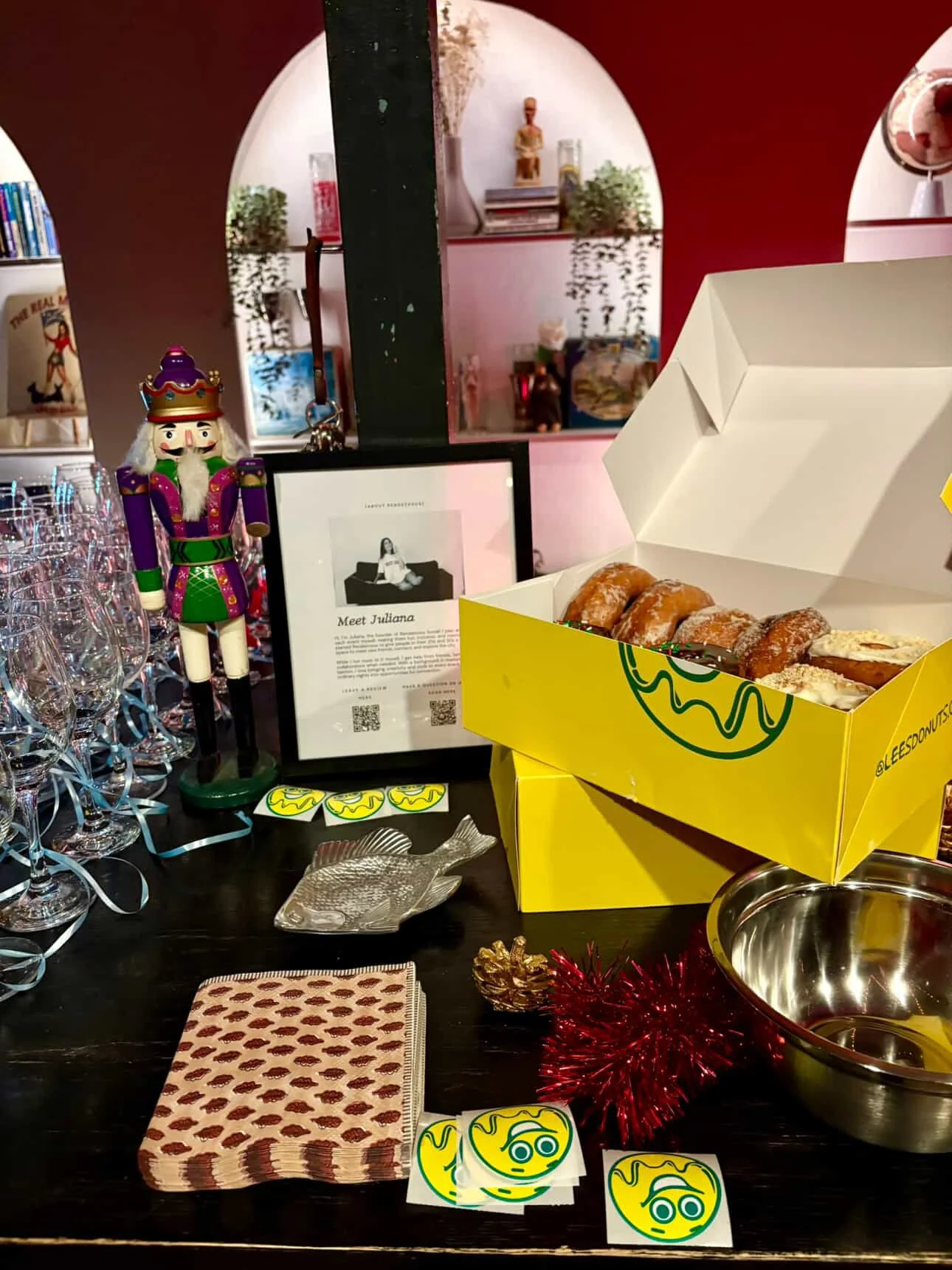 A tabletop decorated with a nutcracker doll, multiple glasses, plates, and Donut boxes, with a framed meet Juliana poster, and shiny holiday decorations.