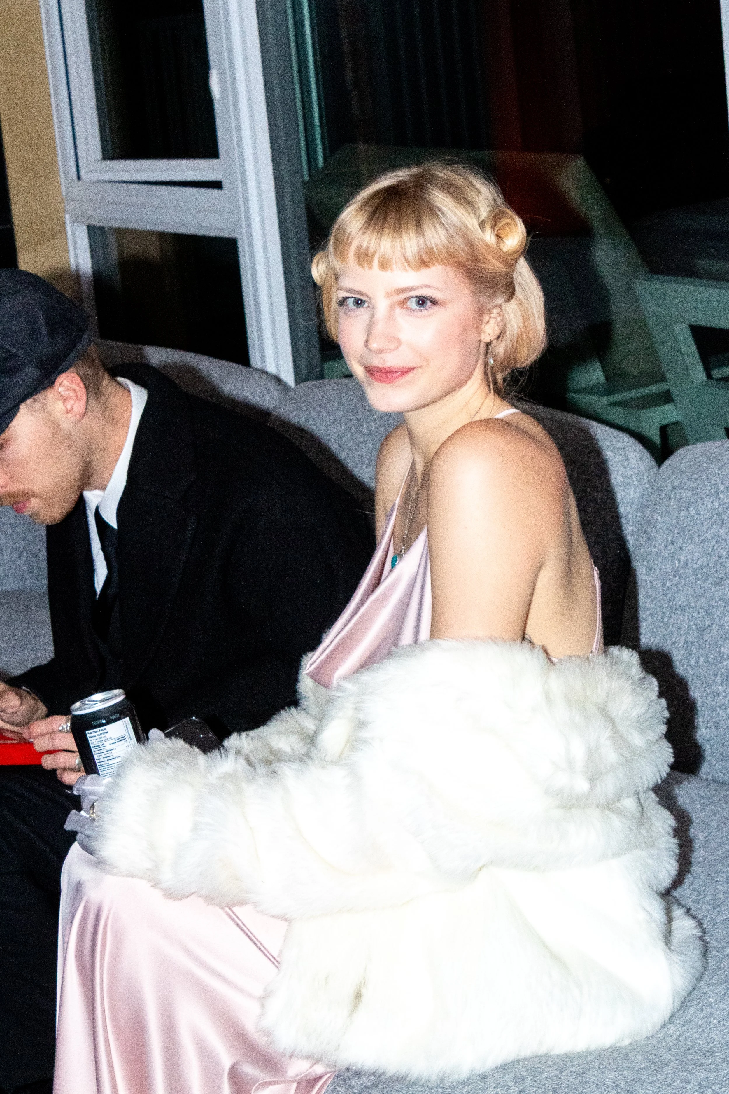 A young woman with blonde hair in vintage style, wearing a satin pink dress and a white fur coat, sitting on a gray sofa.