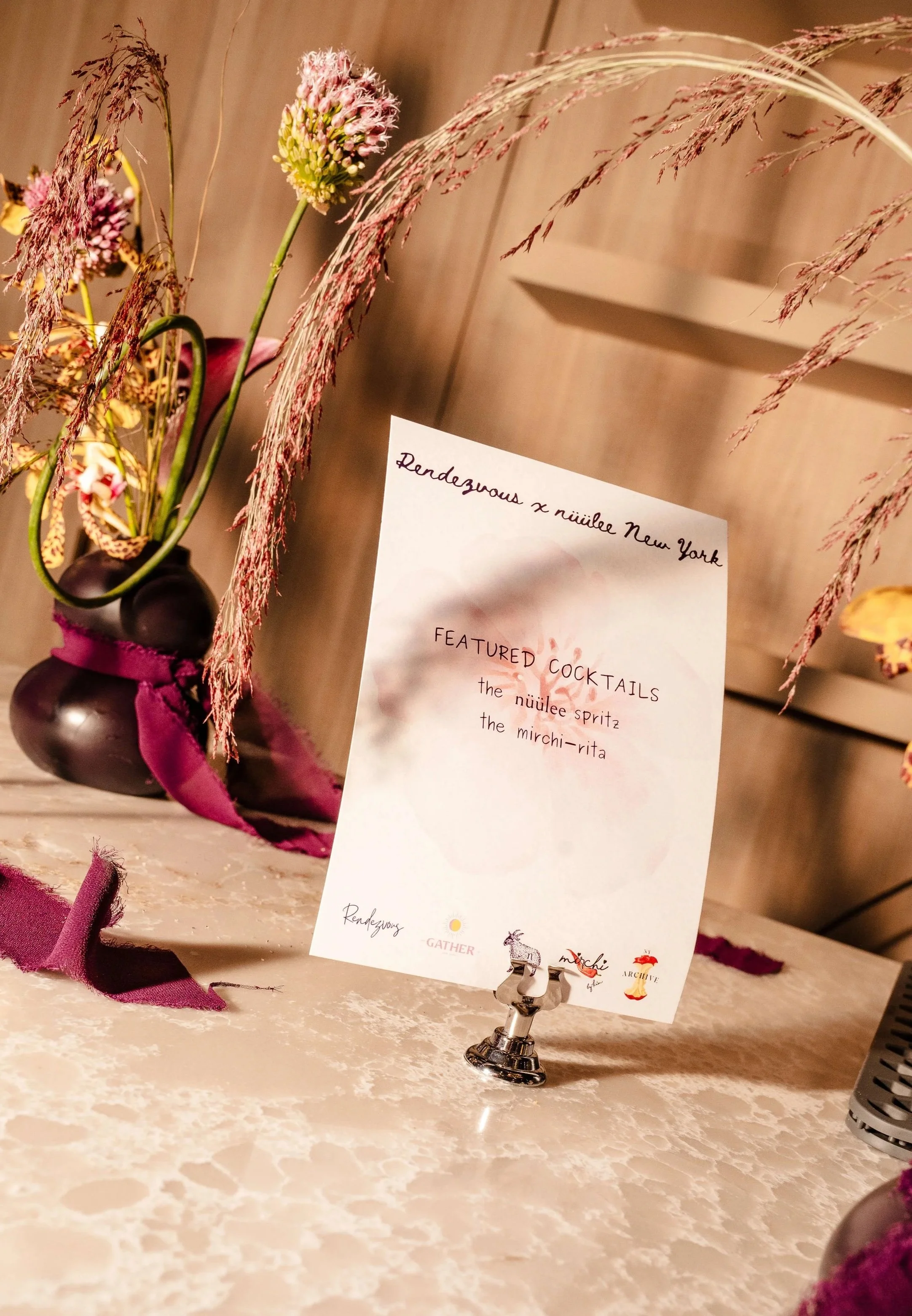 A menu card on a table displays featured cocktails, including the nüülee spritz and the mirchi-rita, with decorative flowers and greenery in the background.