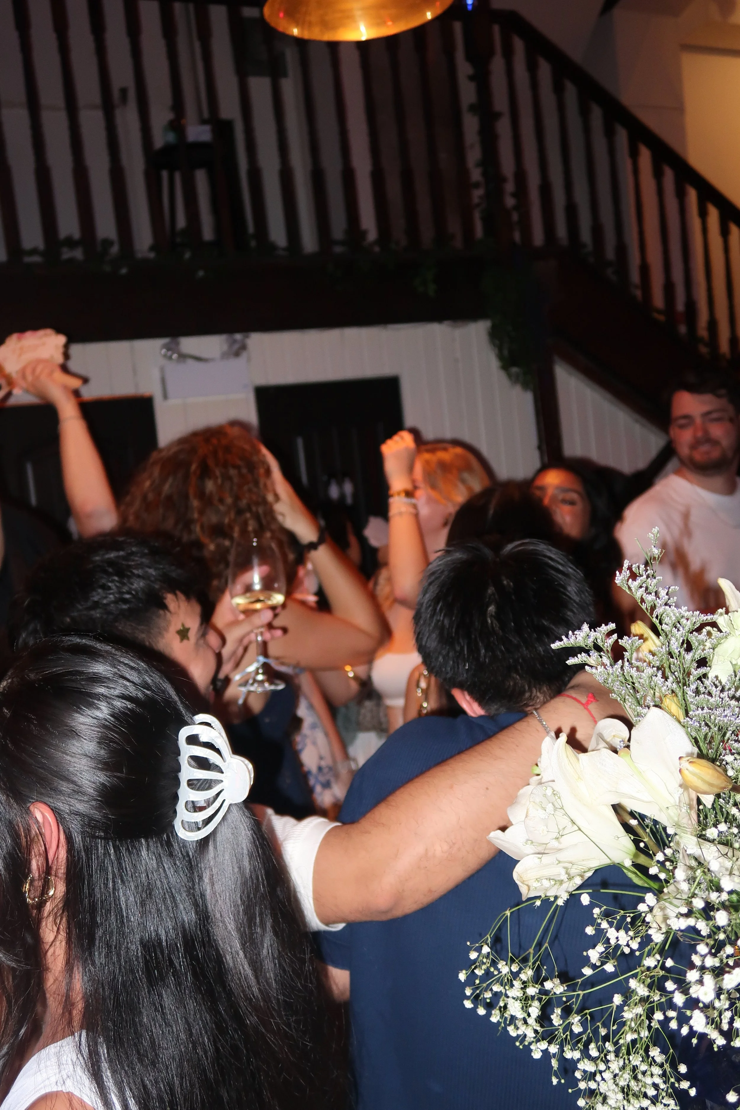Group of people celebrating at a party, with some holding glasses of wine, in a dimly lit indoor setting.