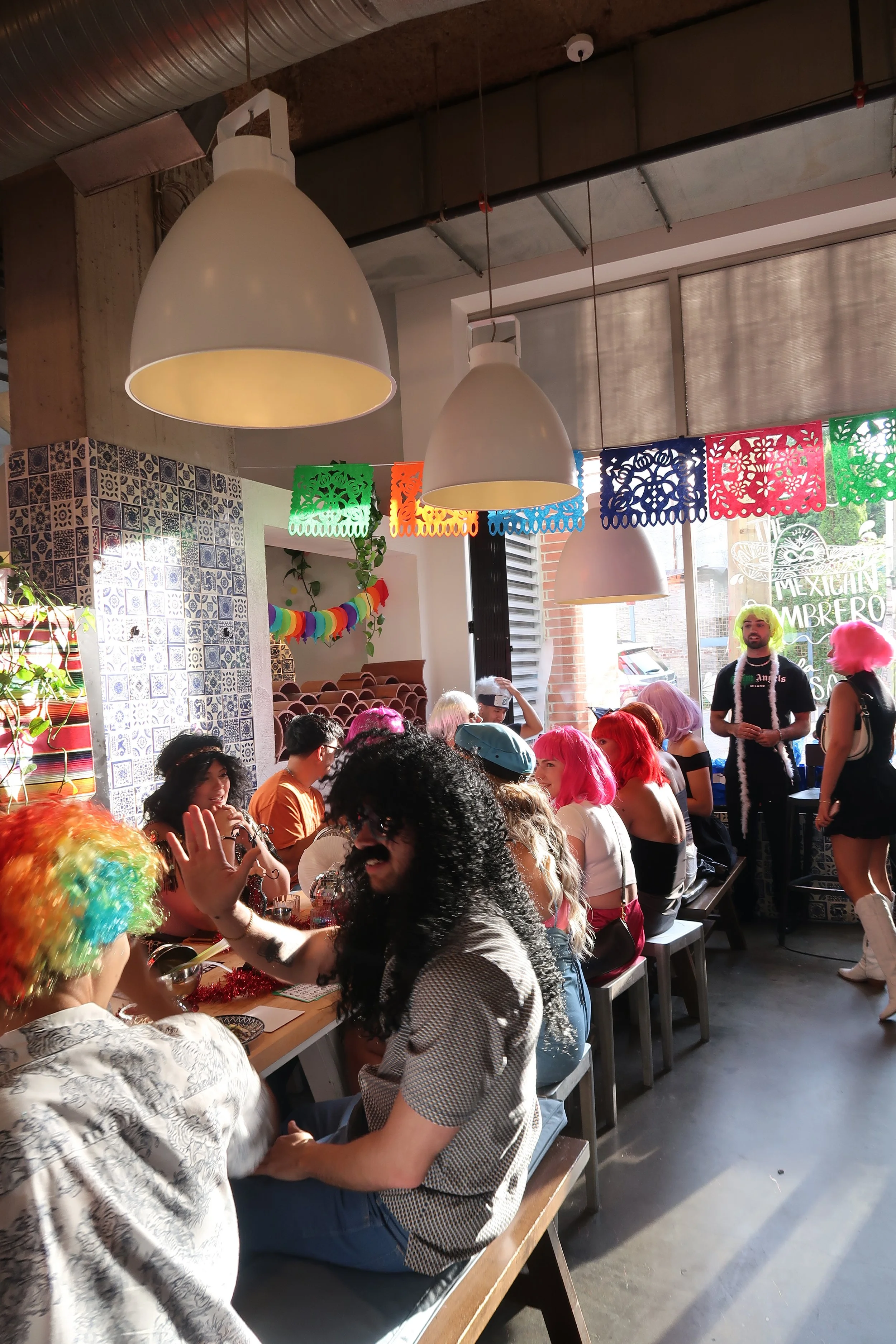 Group of people celebrating a Mexican-themed event at a restaurant, dressed in colorful costumes and wigs, with festive decorations including papel picado banners and a rainbow garland.
