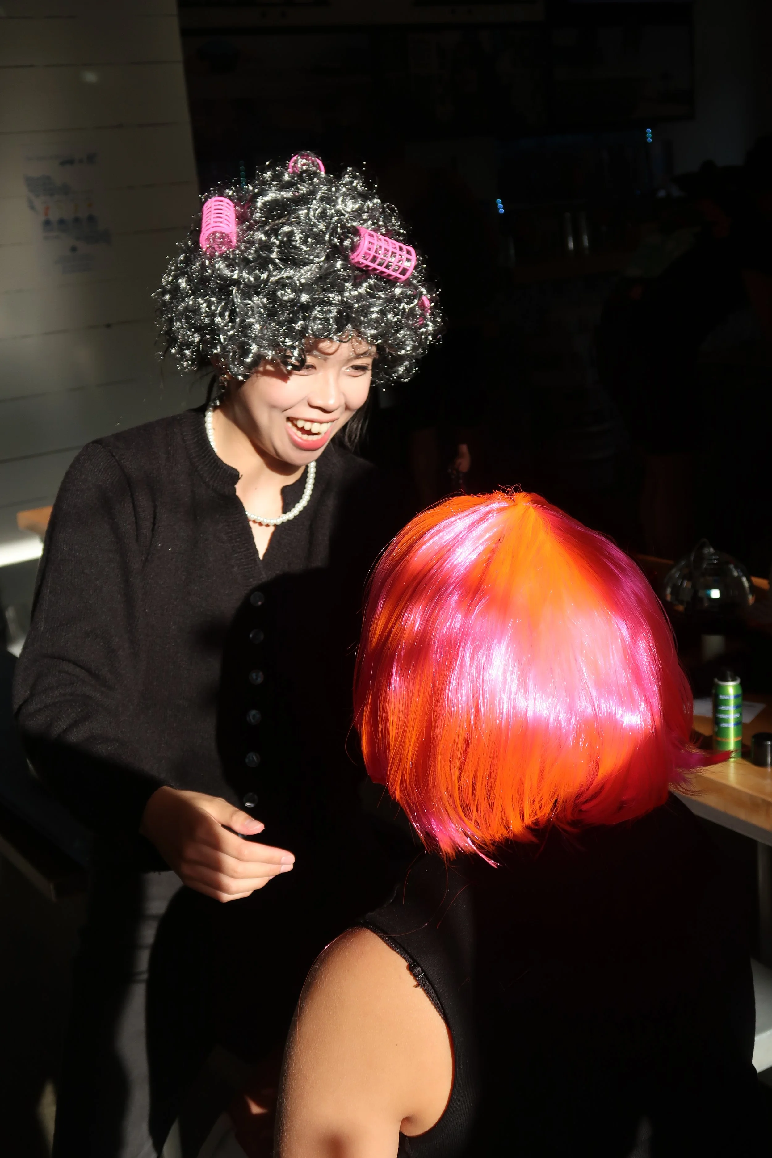 A woman with a joyful expression, wearing a black sweater and pearl necklace, has gray hair rollers and pink curlers in her gray hair. She is facing a person with a bright orange and pink wig, who is seated with their back to the camera, in a dimly l