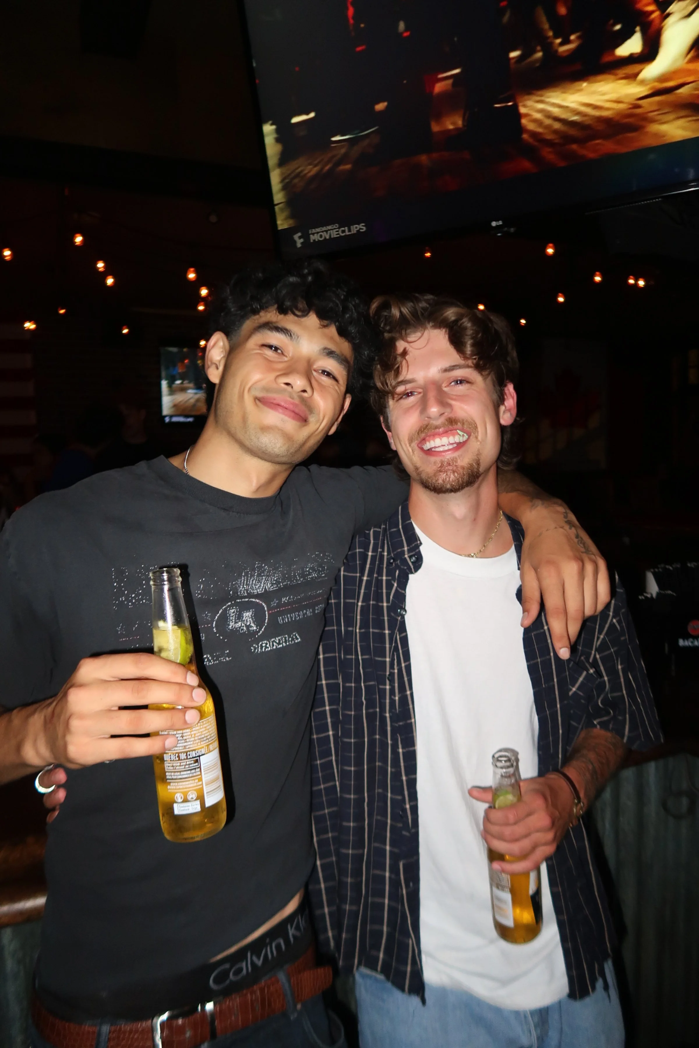 Two young men smiling and holding beer bottles at a bar, with dim lighting and string lights overhead.