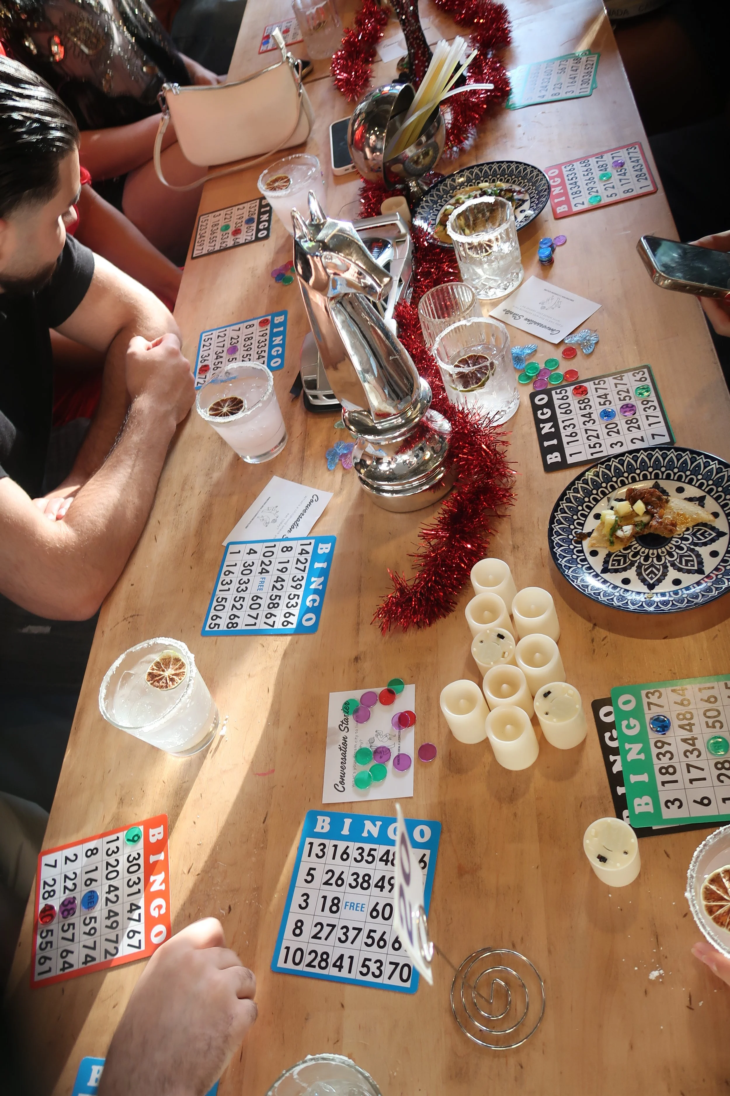 A table decorated with red tinsel and multiple bingo cards, glasses with drinks and lemon slices, candles, and party favors during a game of bingo with several people gathered around.
