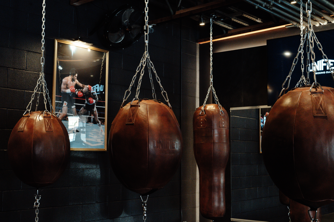 Inside a boxing gym with hanging leather punching bags, a framed picture of a boxer in action on a black brick wall, and a large digital screen showing a boxing match.