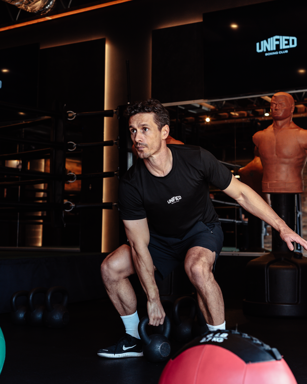 A man lifting a kettlebell during a workout at a modern gym, with punching bags and a sign reading 'United Boxing Club' in the background.