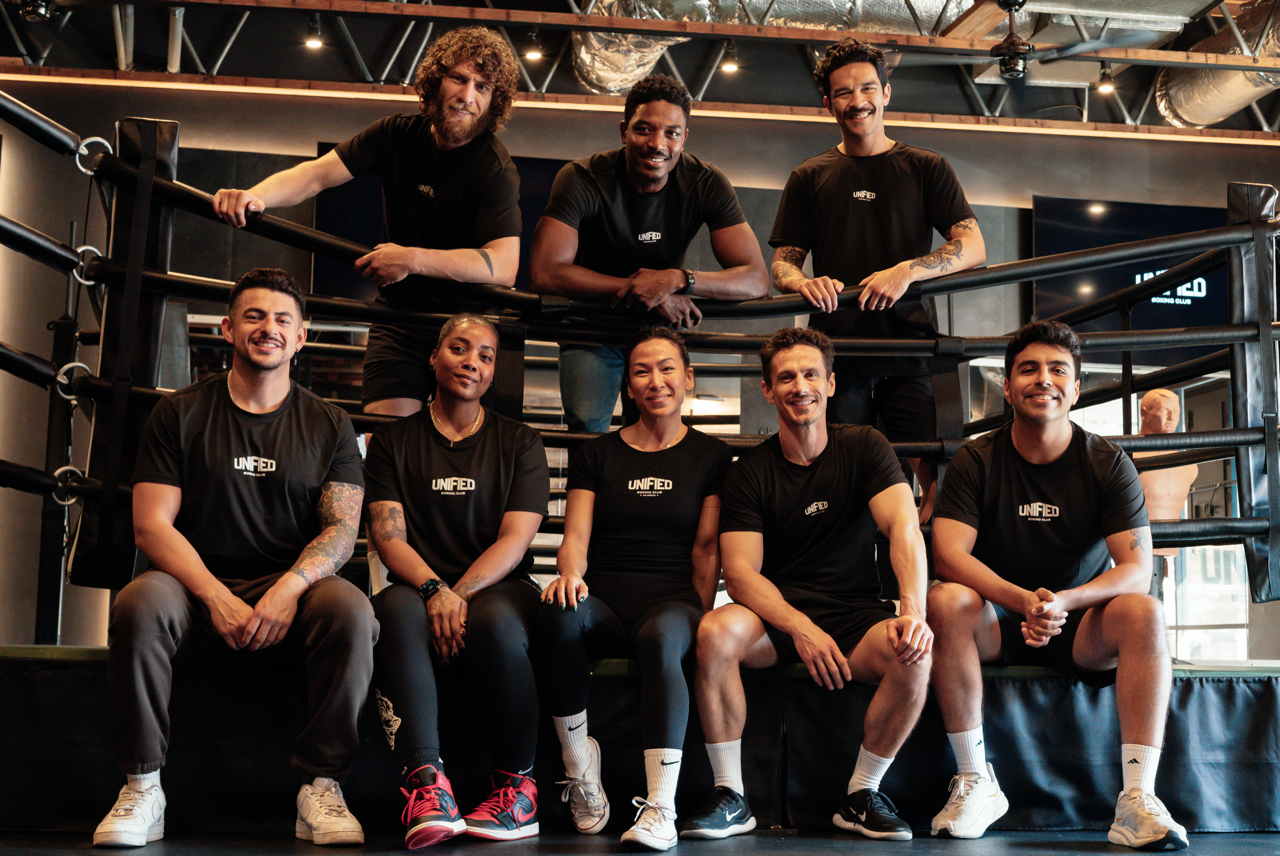 A group of nine diverse people in black T-shirts with 'UNIFIED' written on them, posing inside a boxing gym near a boxing ring, smiling at the camera.