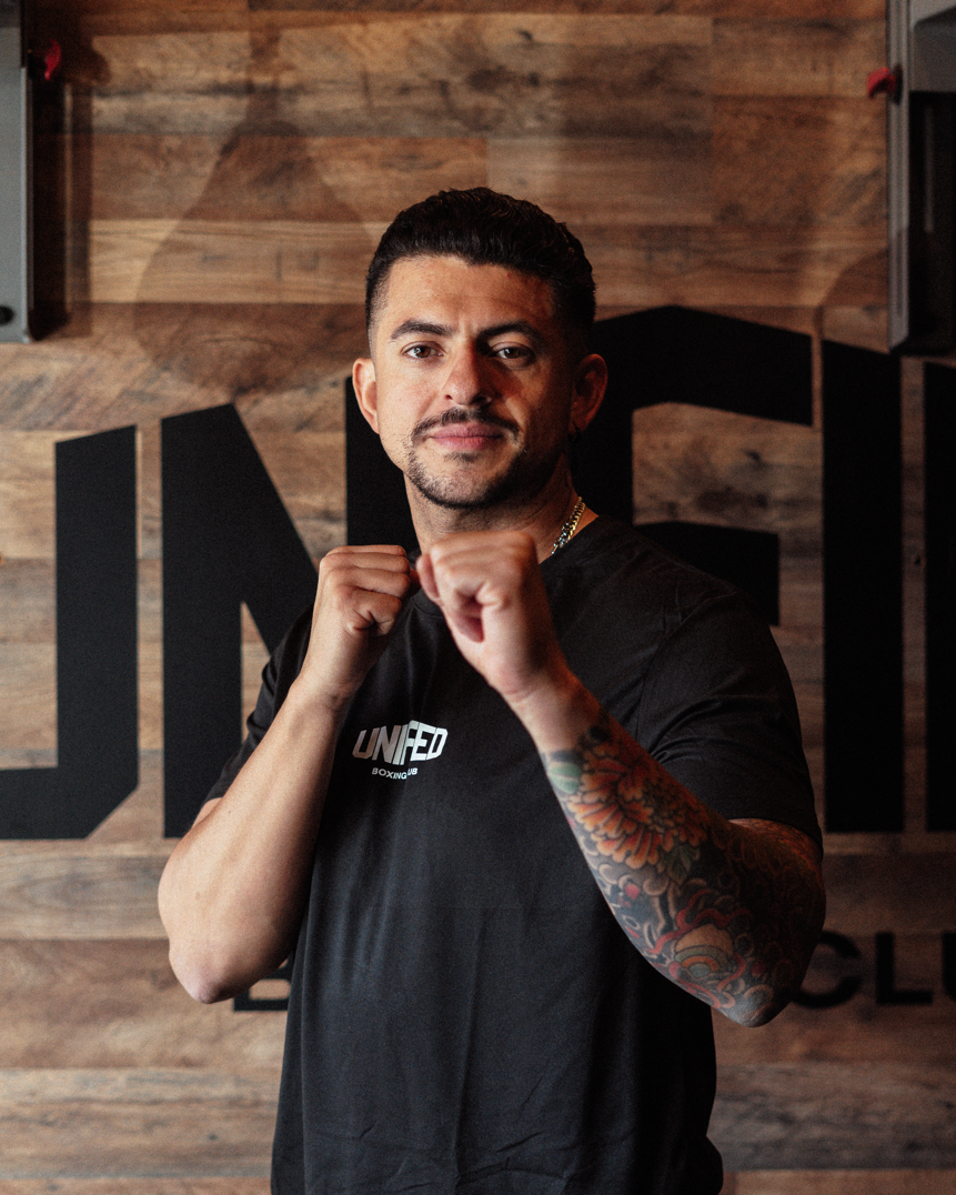 A man with a baseball cap, earrings, and tattooed arm leaning on a railing, speaking with an expressive face, inside a venue with a screen in the background displaying the word 'United'.