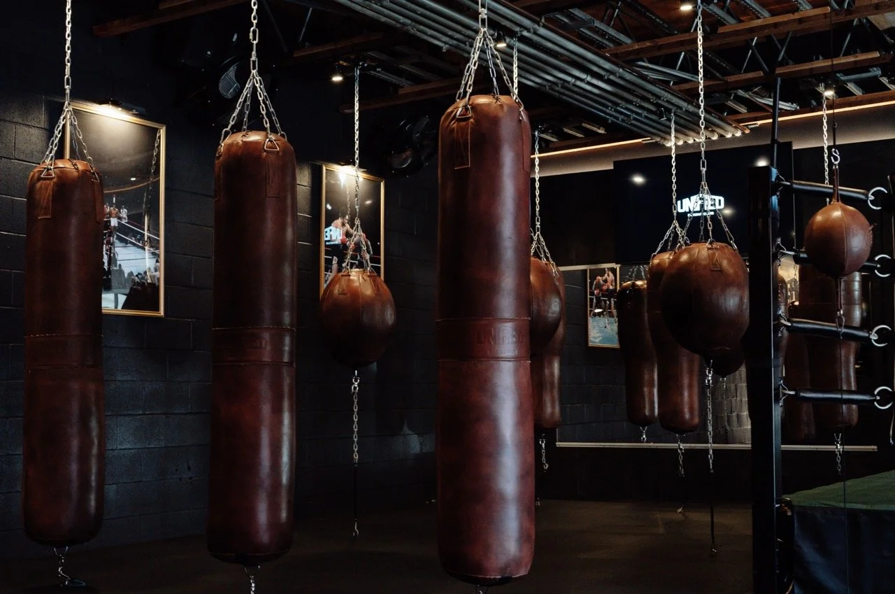 Multiple brown leather punching bags hanging in a gym or boxing studio, with black walls and framed photos on the wall.