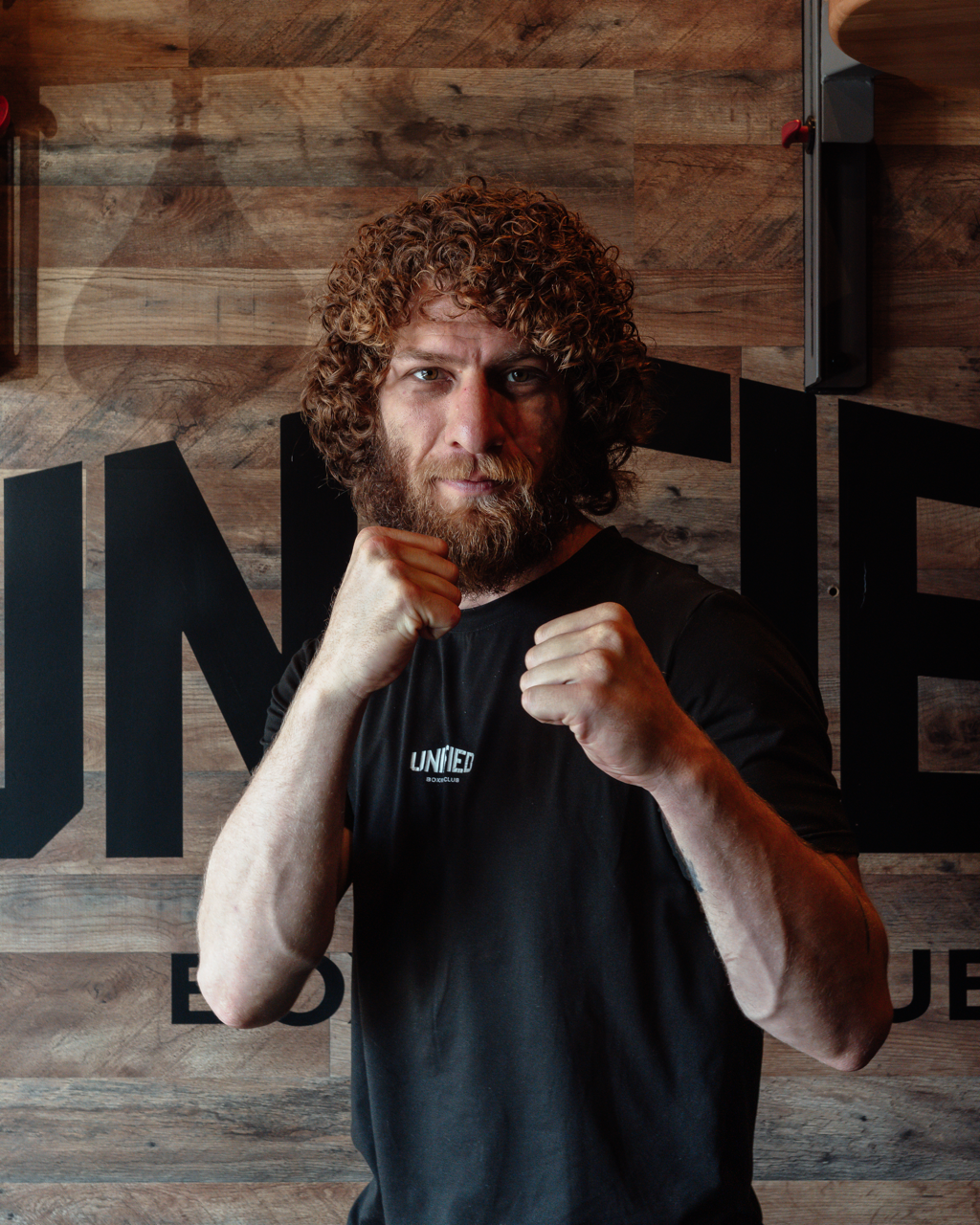 A group of people, including a man with curly hair and a beard, are in a boxing gym preparing for a session. They are standing near punching bags, with some wrapped in cloth or tape, and are listening to instructions.