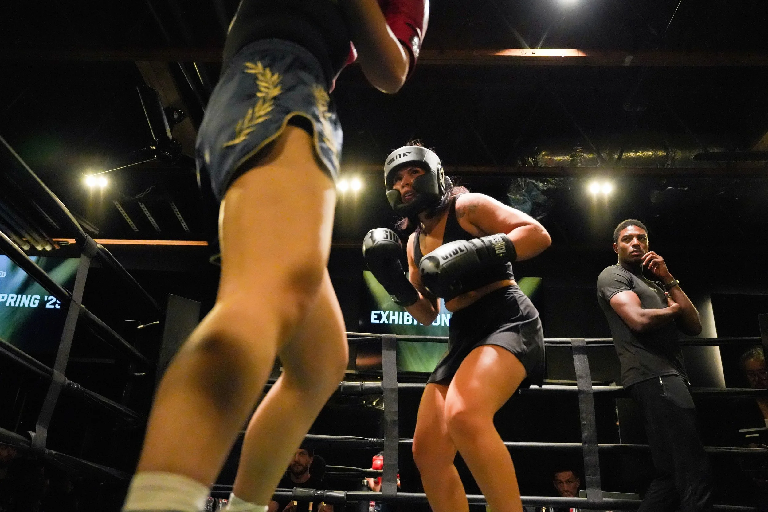 Female boxer wearing a helmet and gloves practicing in a boxing ring, with a coach or referee observing nearby.