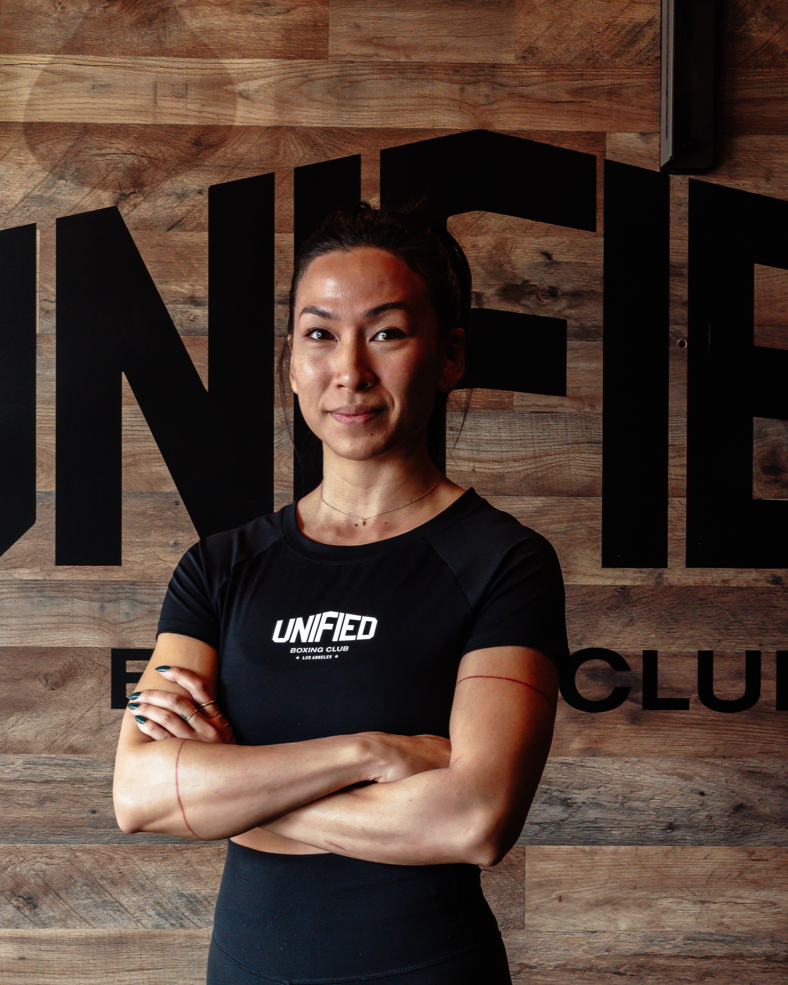 Woman taking boxing or kickboxing class, holding focus mitts against a male instructor who is throwing a punch. Other people practicing in the background inside a gym with wooden walls and large windows.
