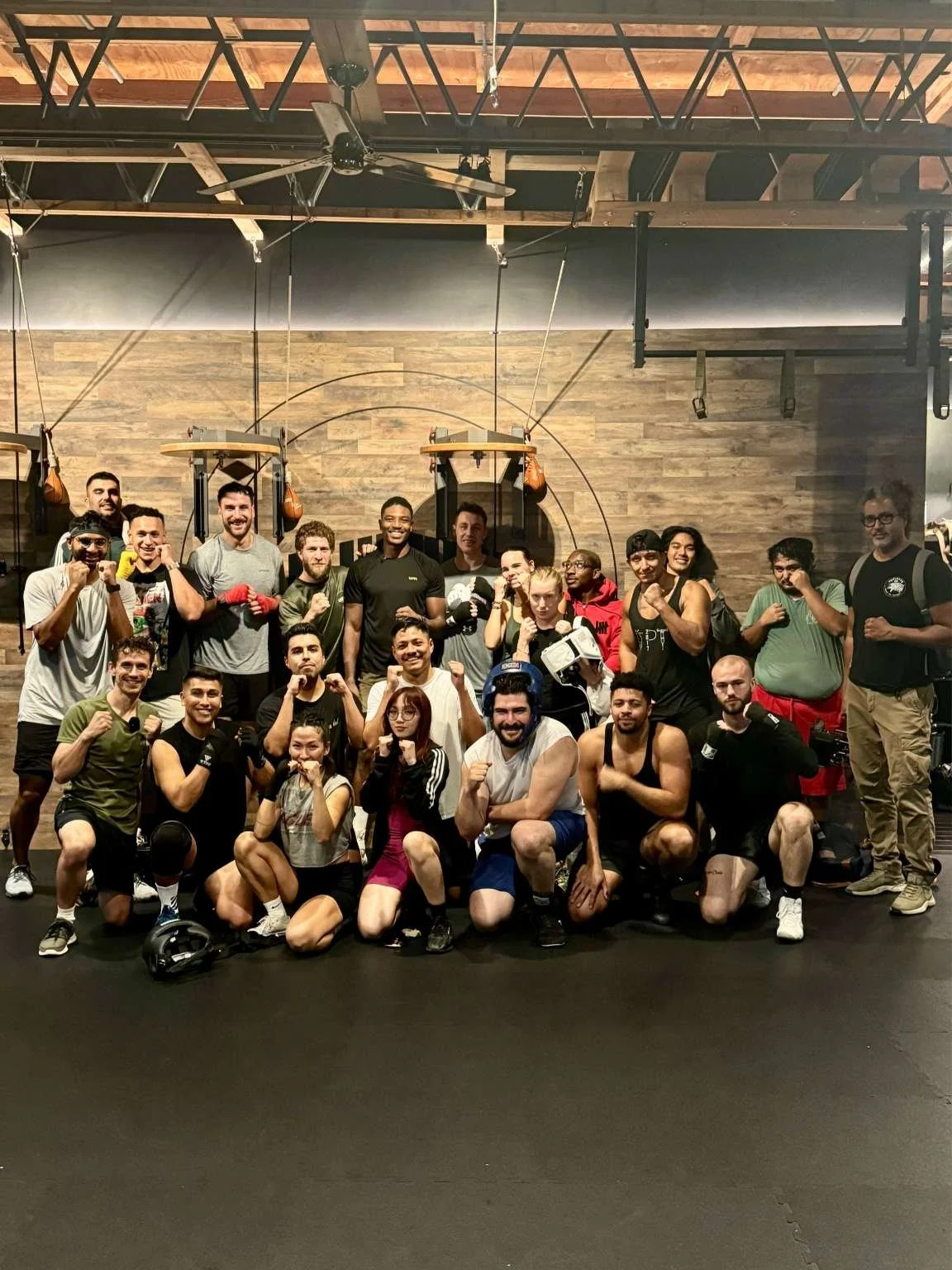 Group of diverse people in a martial arts gym, some with boxing gloves, posing with fists raised, several people in kneeling positions, with a wooden wall and fitness equipment in the background.