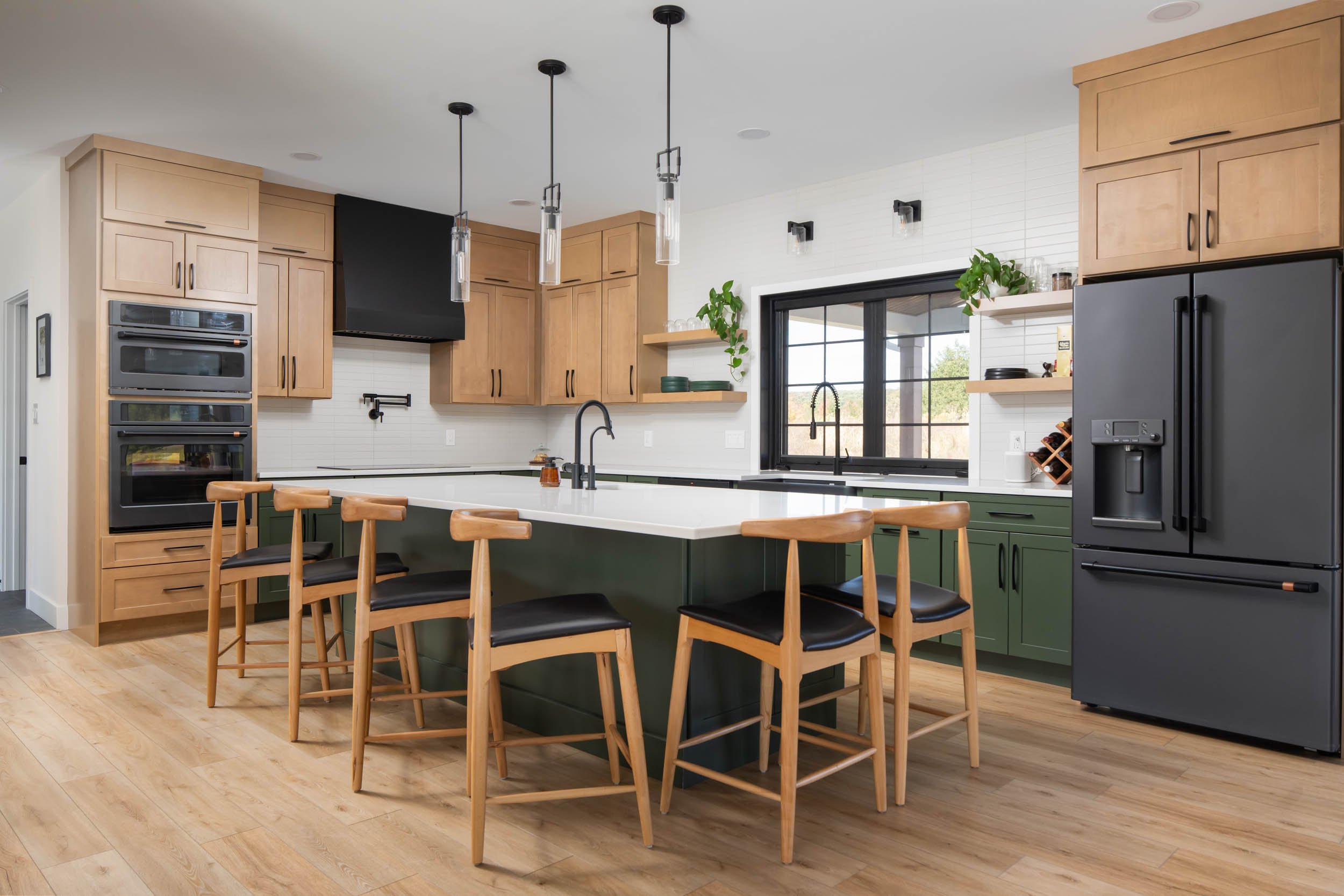 Modern kitchen with natural wood, black, and green cabinets, a large island with a white countertop, wooden chairs with black seats, black appliances, and pendant and wall-mounted lighting fixtures.