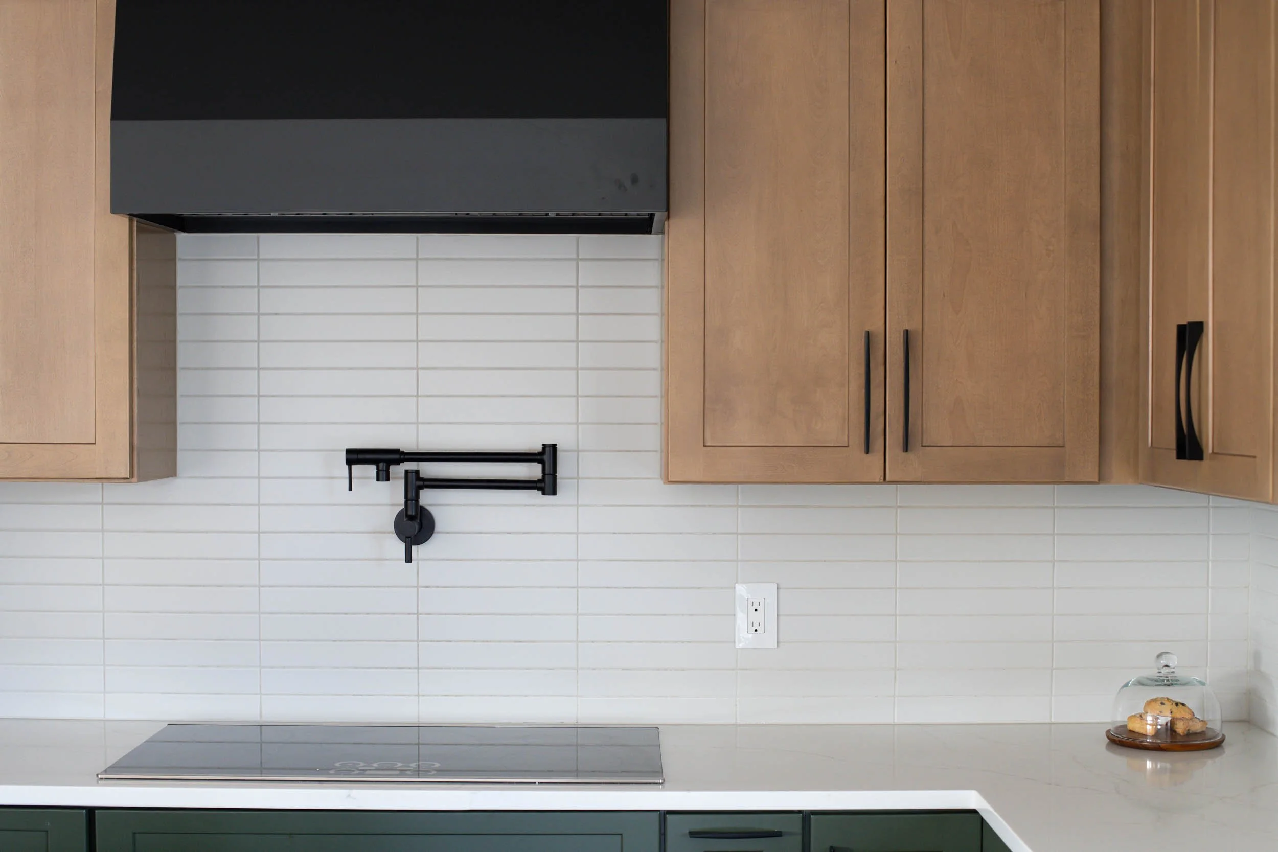 Modern kitchen with brown cabinets, white subway tile backsplash, black range hood, black faucet, and a white countertop with a glass cake dome and cookies.