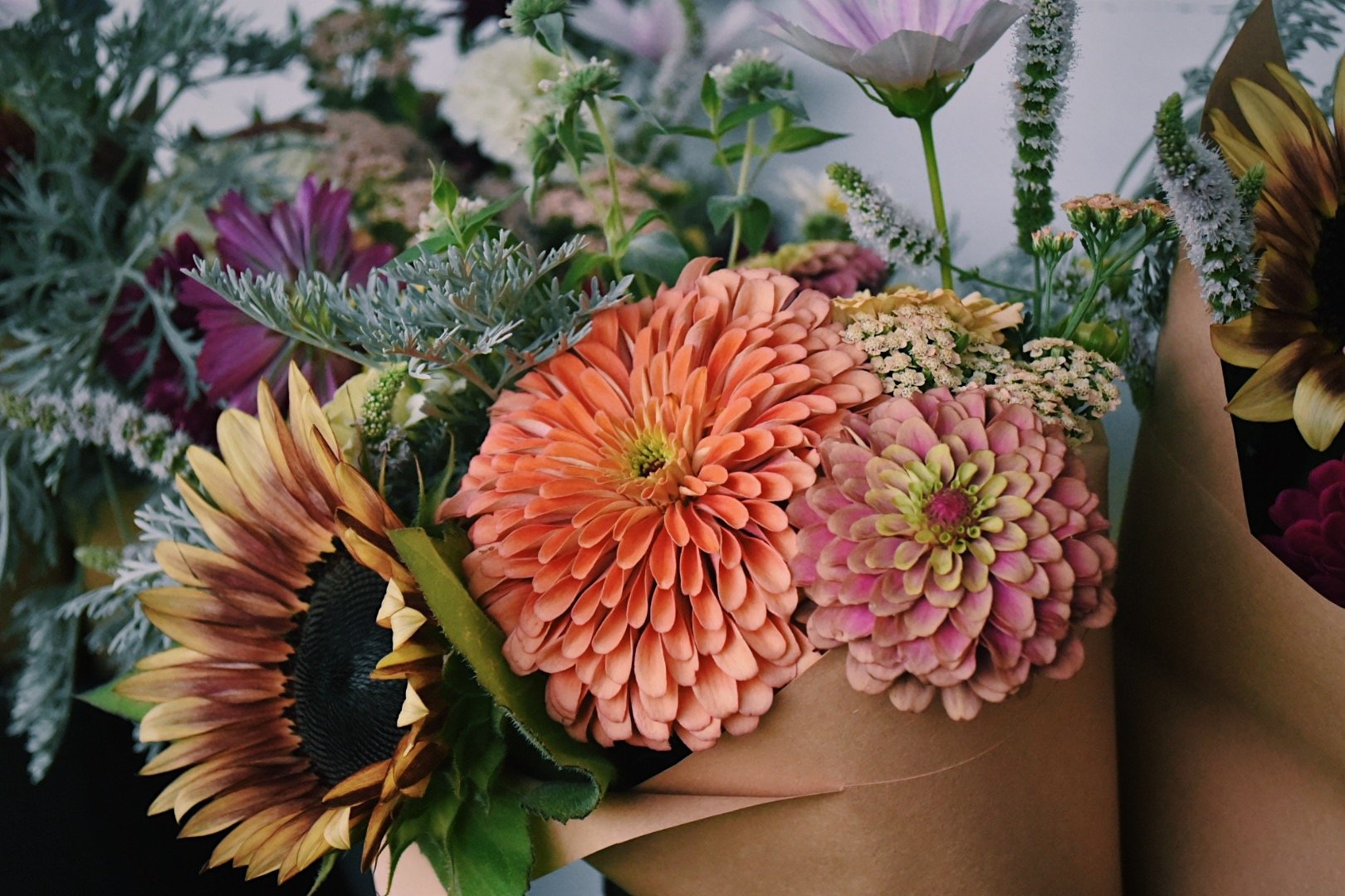 A bouquet of colorful flowers including sunflowers, zinnias, daisies, and various greenery wrapped in brown paper.