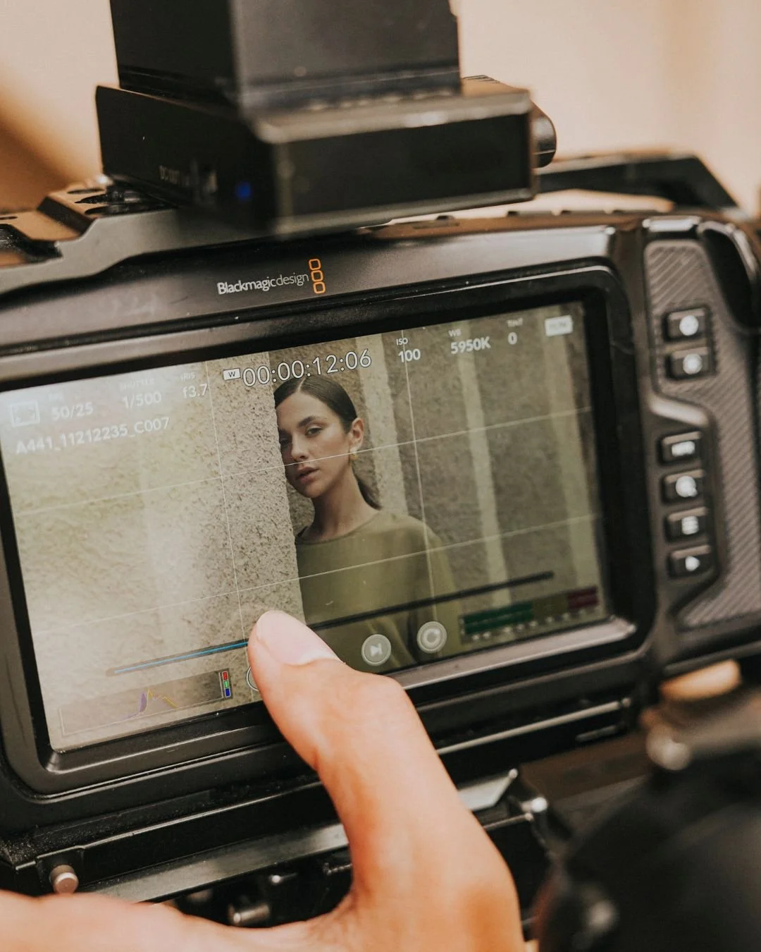 A camera viewfinder captures a woman standing against a textured beige wall, with a focus on her face and shoulder, as a person presses a button on the camera.
