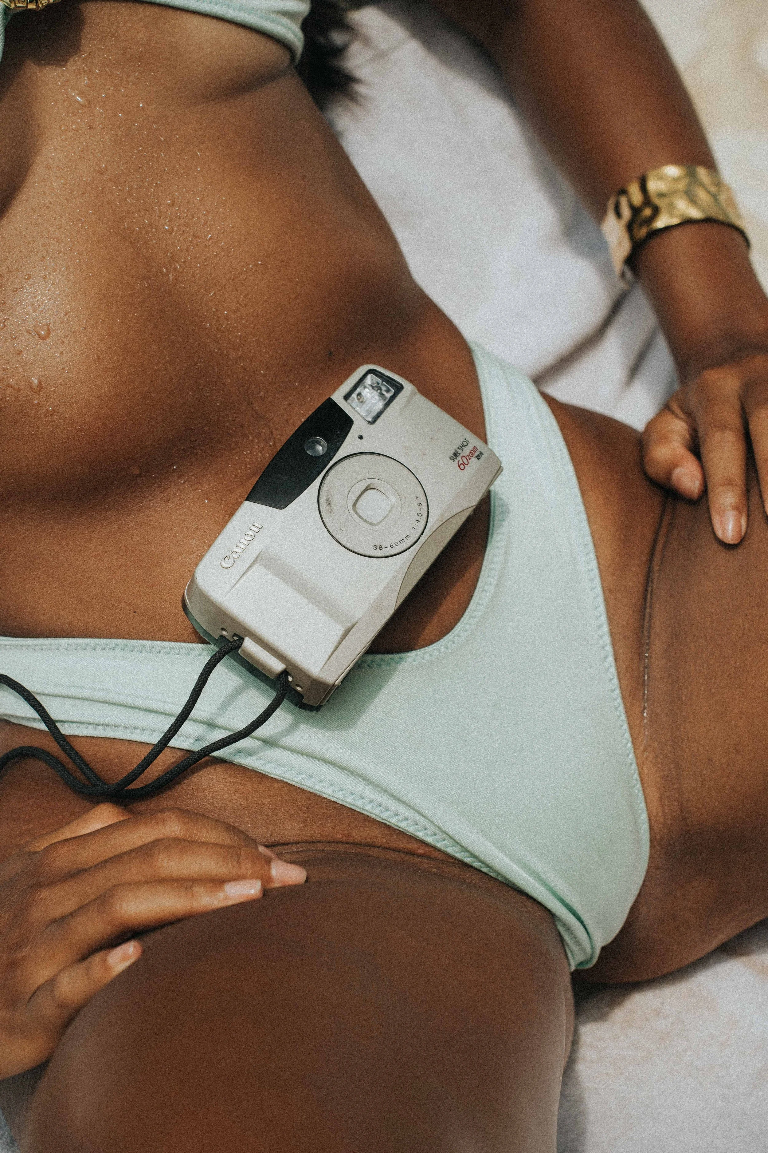 Close-up of a woman lying down on a white surface with a camera placed on her waist, wearing lavender underwear and a gold bracelet.