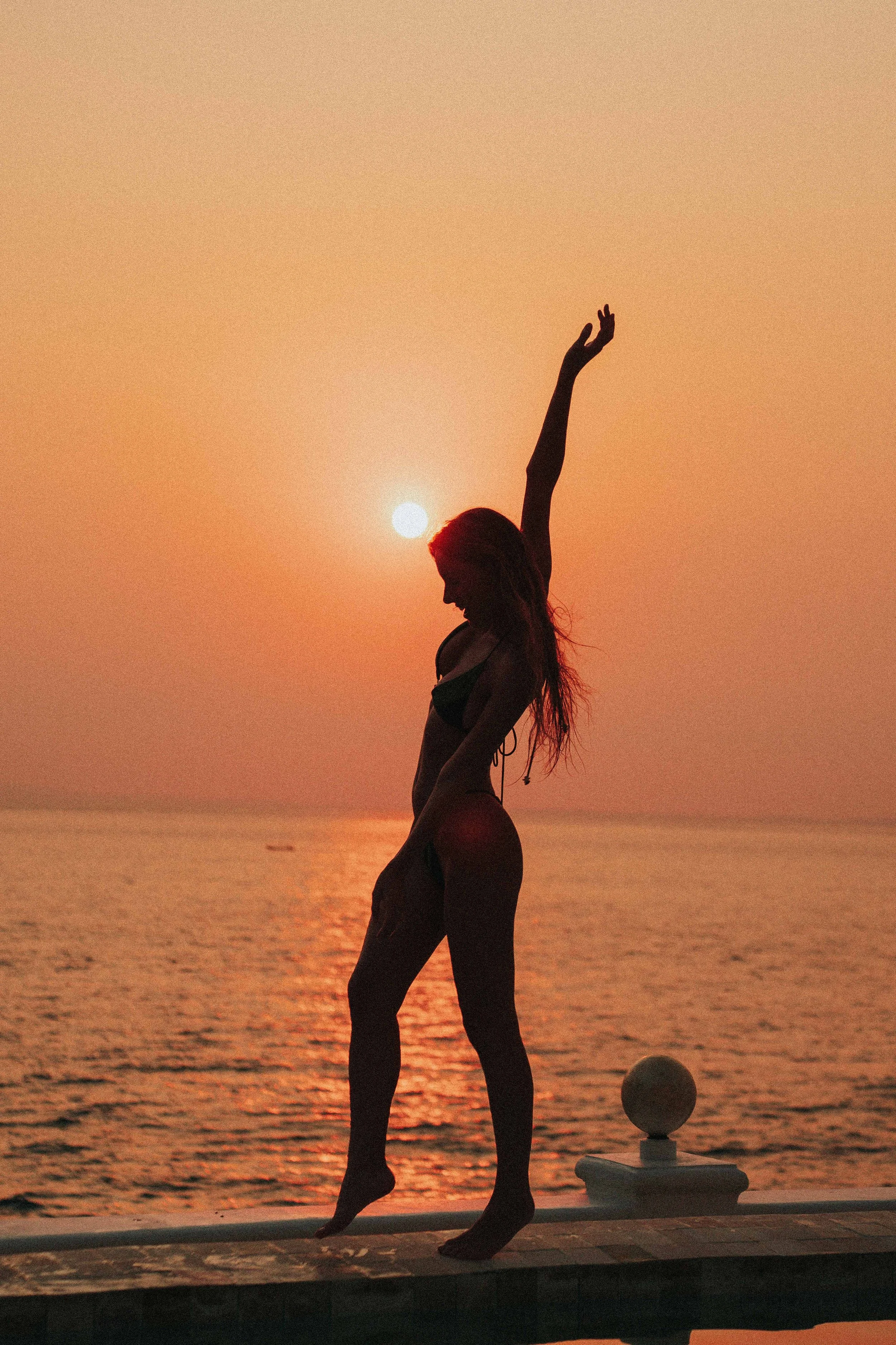 Silhouette of a woman in a bikini standing on a ledge at sunset or sunrise near the water, with one arm raised above her head and the sun in the background.