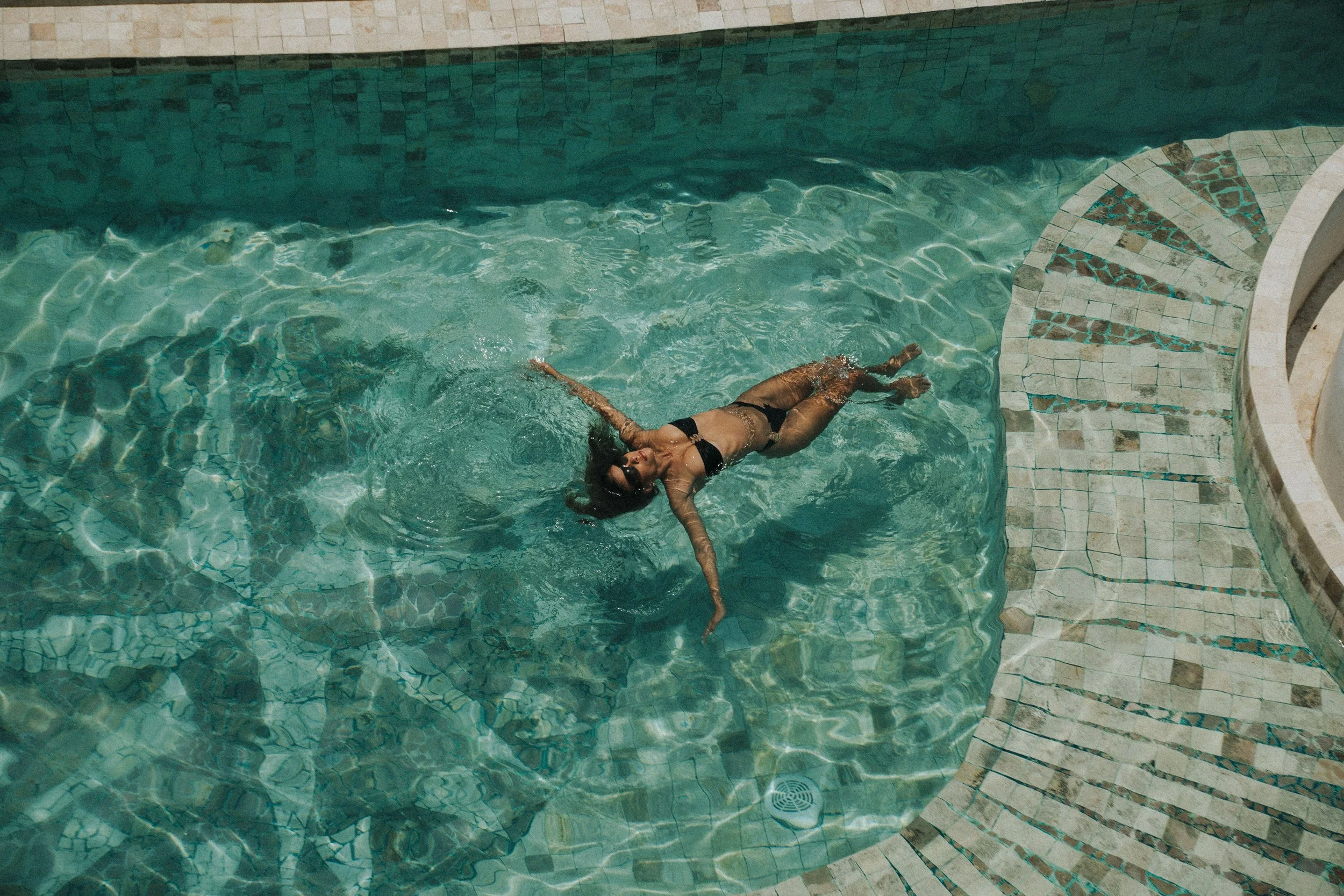 Woman in a black bikini and sunglasses floating and relaxing in a swimming pool with tiled steps nearby.