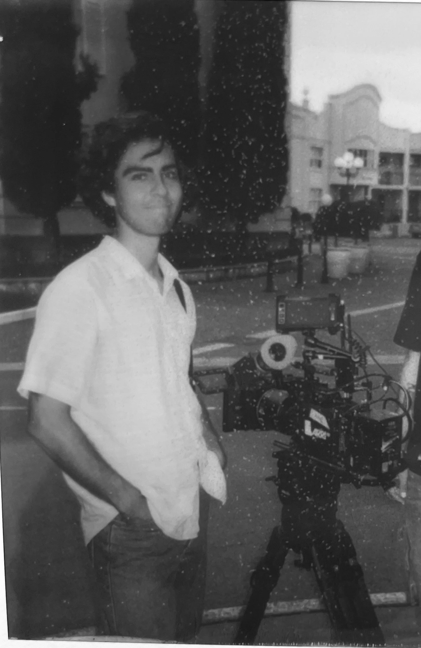 A young man with dark hair and a slight smile, standing outdoors near filming equipment, at dusk or nighttime with a building and trees in the background.