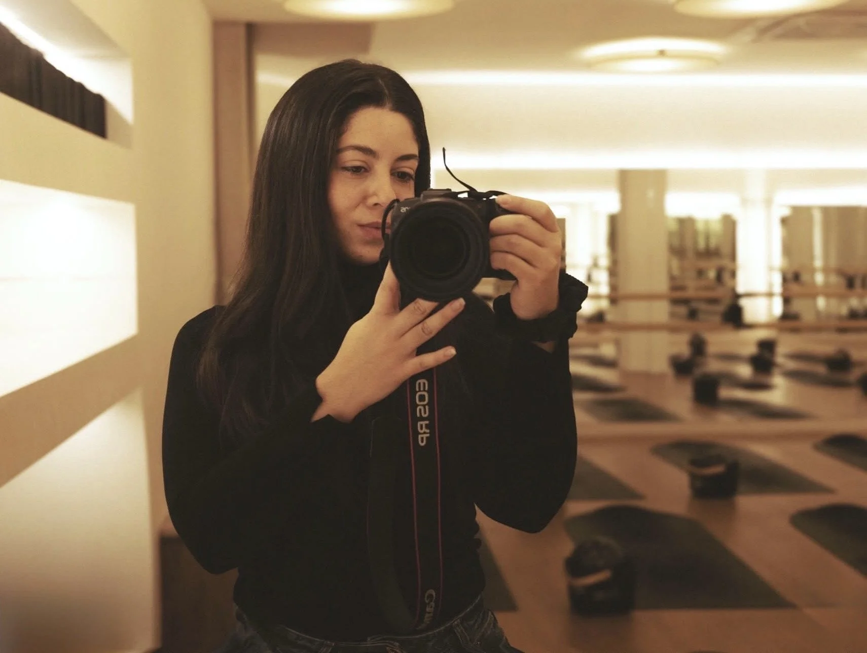 A woman taking a selfie in a mirror with a camera in a dimly lit room that appears to be a gym or wellness center with yoga mats on the floor.