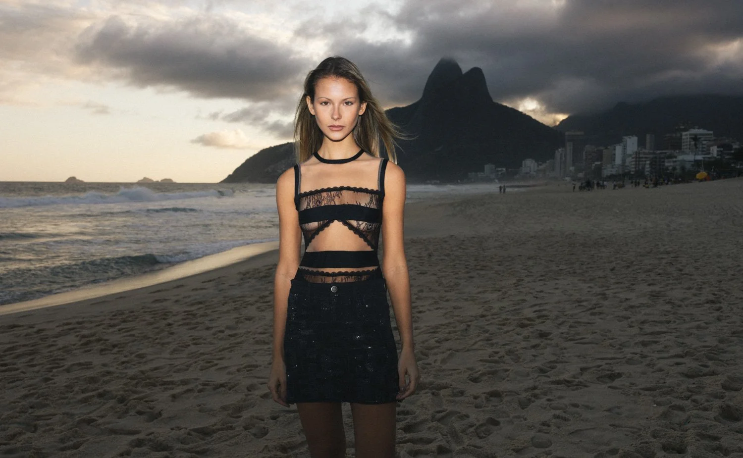 A woman standing on a beach at sunset, wearing a sheer black lace top and a black skirt, with mountains and a cloudy sky in the background.