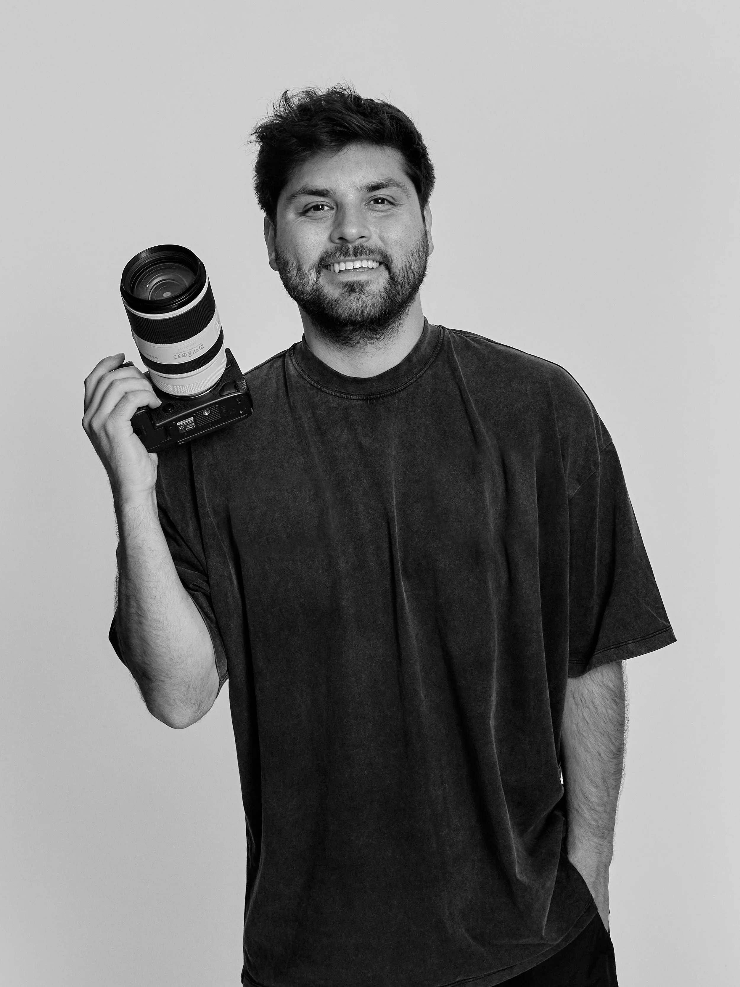 A man with a beard and messy hair smiling while holding a camera with a large lens over his shoulder, against a plain light-colored background.