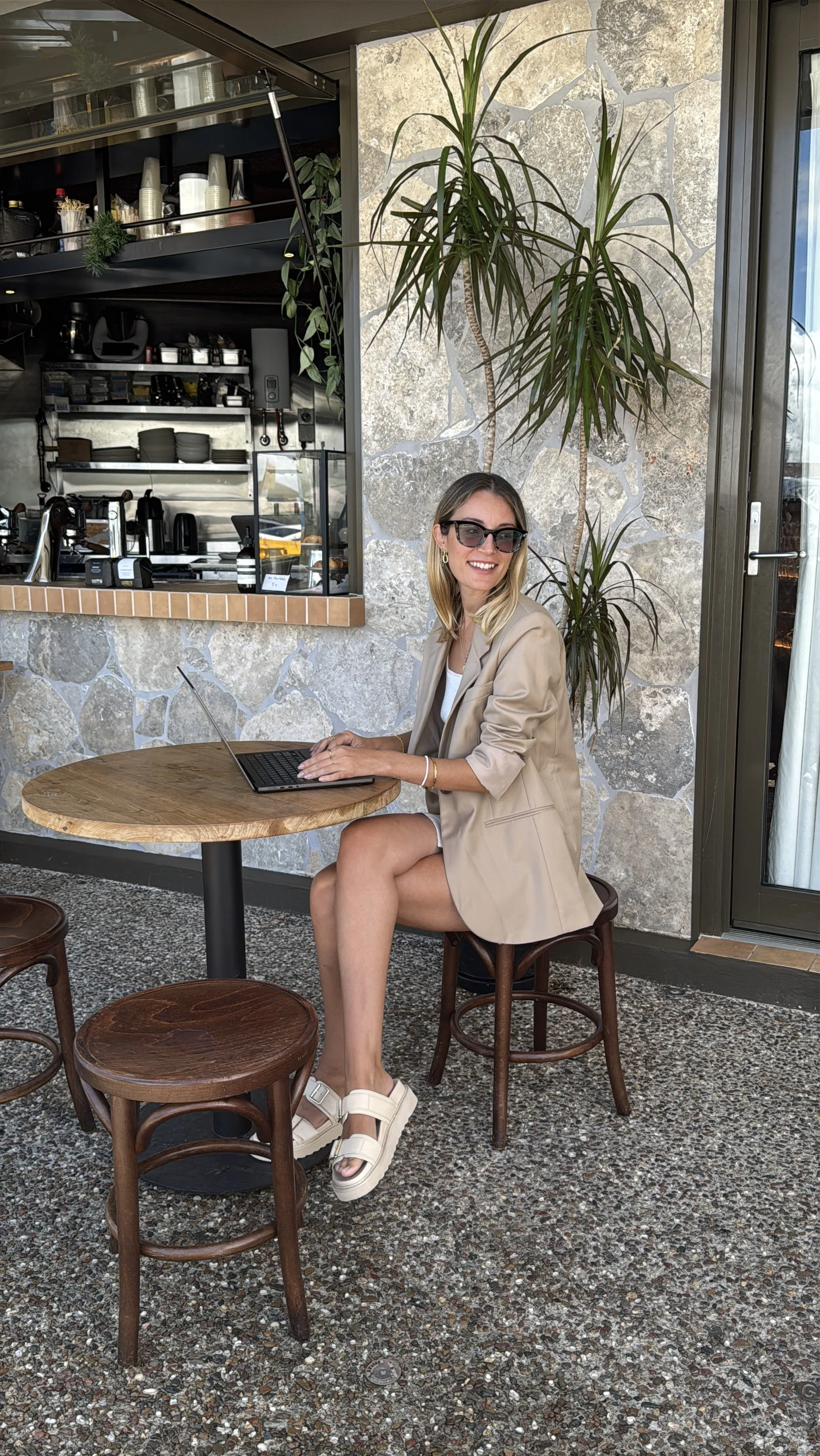 A woman with blonde hair, wearing sunglasses, a beige blazer, white dress, and white platform sandals, sitting at a round wooden table with a laptop, in a cafe with a stone wall and green plants in the background.