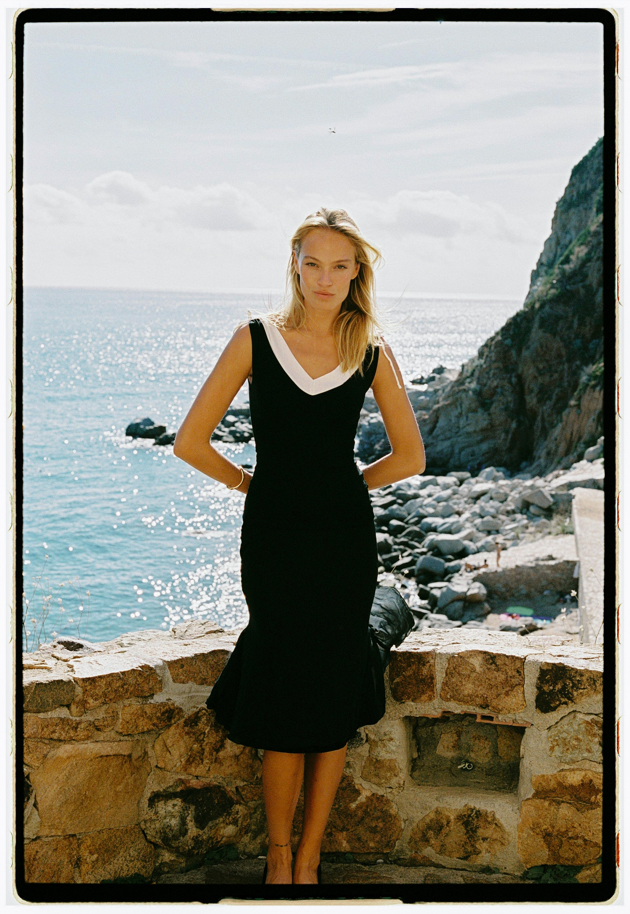 A woman in a black dress with white trim stands on a stone wall by the water, with rocks and a cliff behind her, near a sunny coastline.