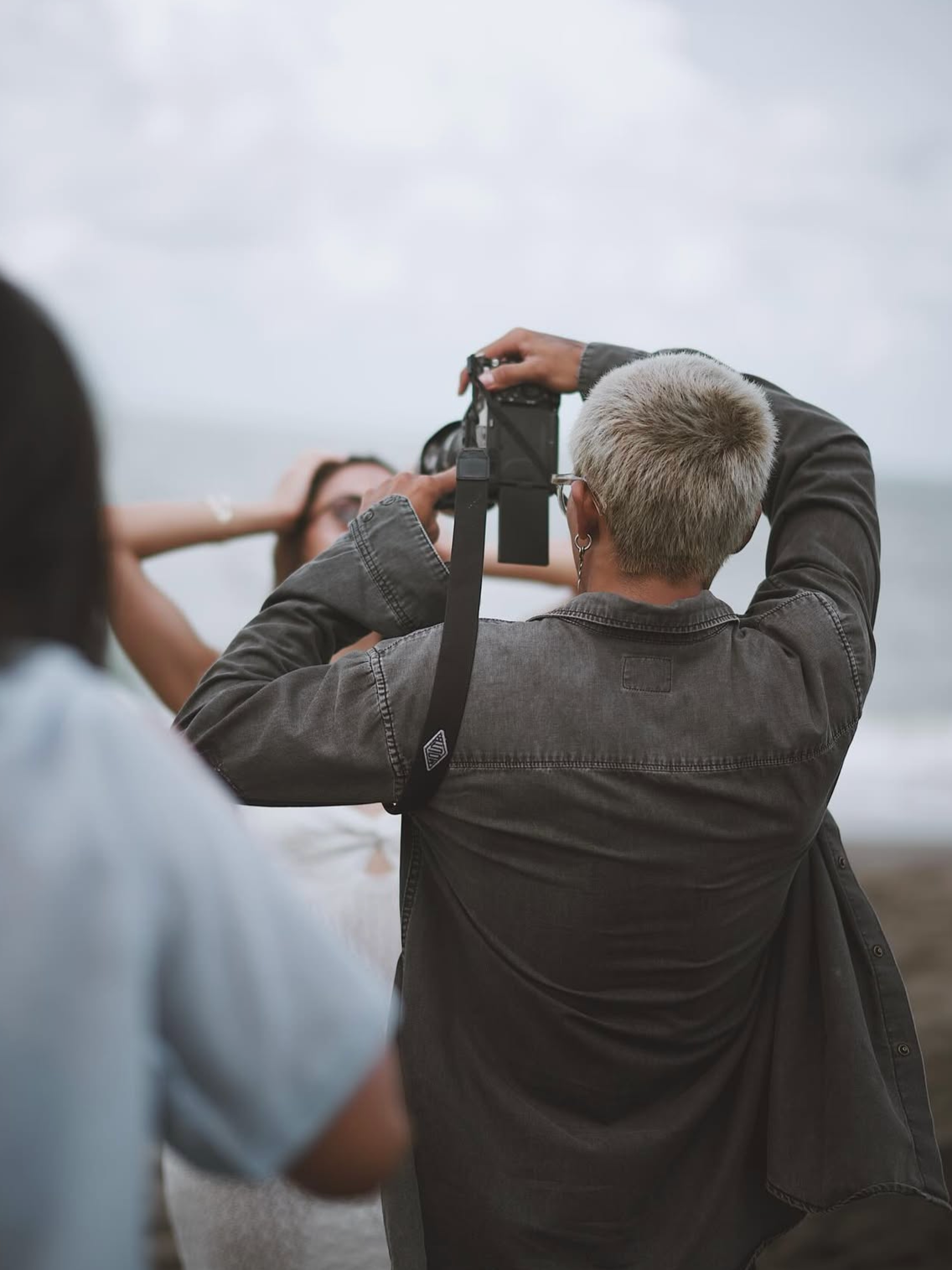 Person with short, gray hair taking a photo of a woman at the beach on a cloudy day.