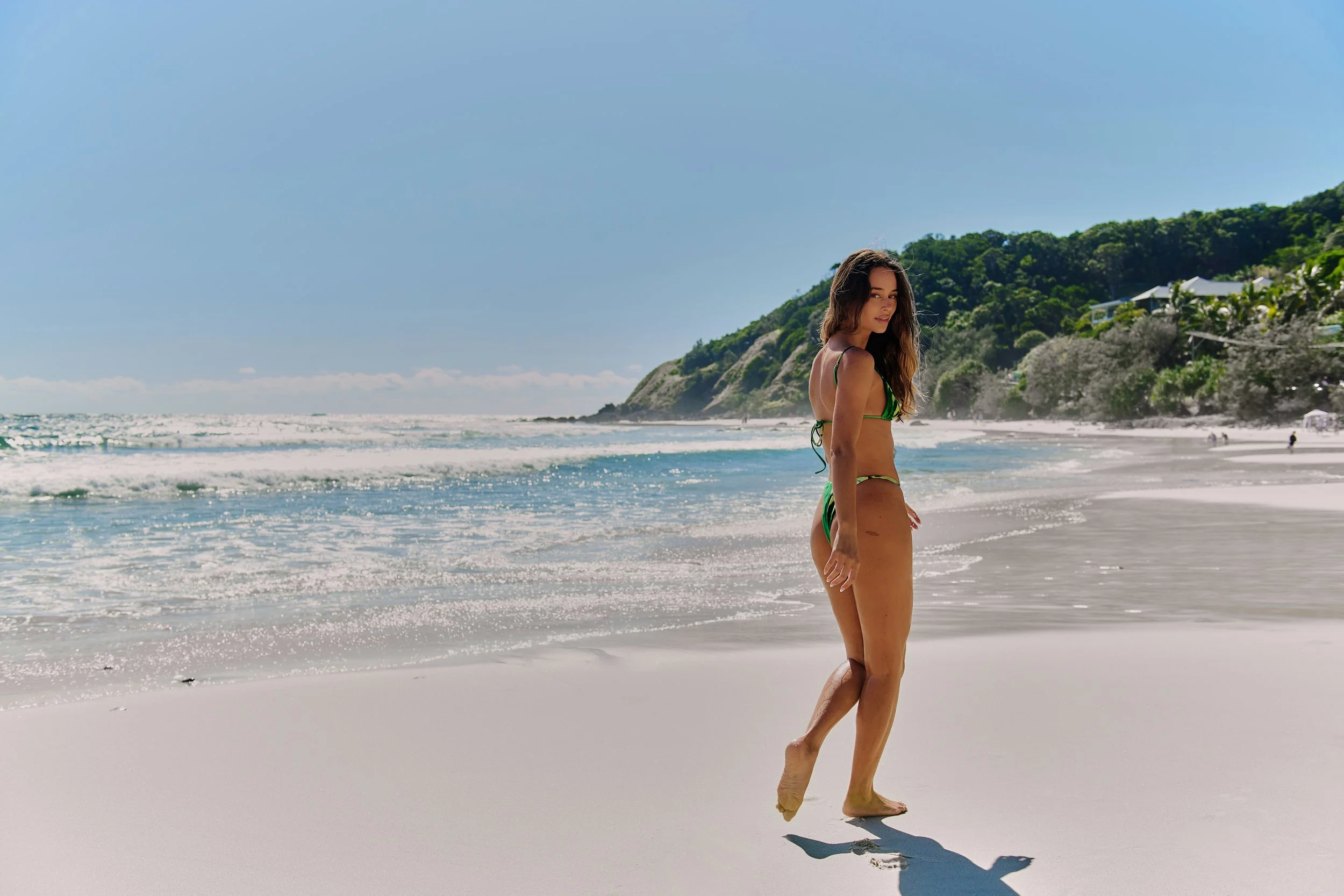 A woman in a green bikini walking along the shoreline of a beach with waves, a green hillside, and houses in the background.