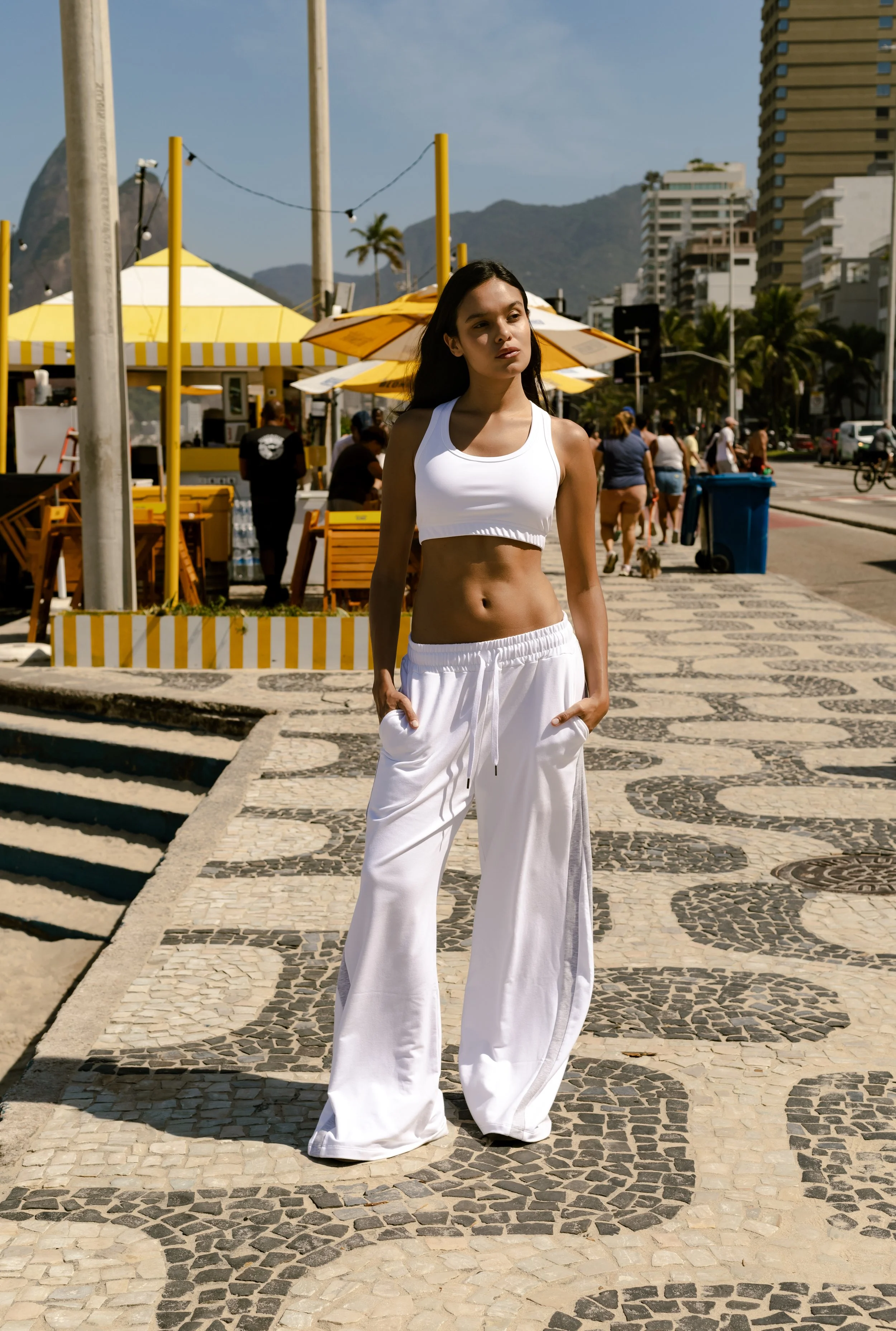 A young woman with long dark hair stands on a paved walkway in a sunny outdoor area, wearing a white crop top and loose white pants, with her hands in her pockets. There are yellow and white striped umbrellas and people walking in the background, wit