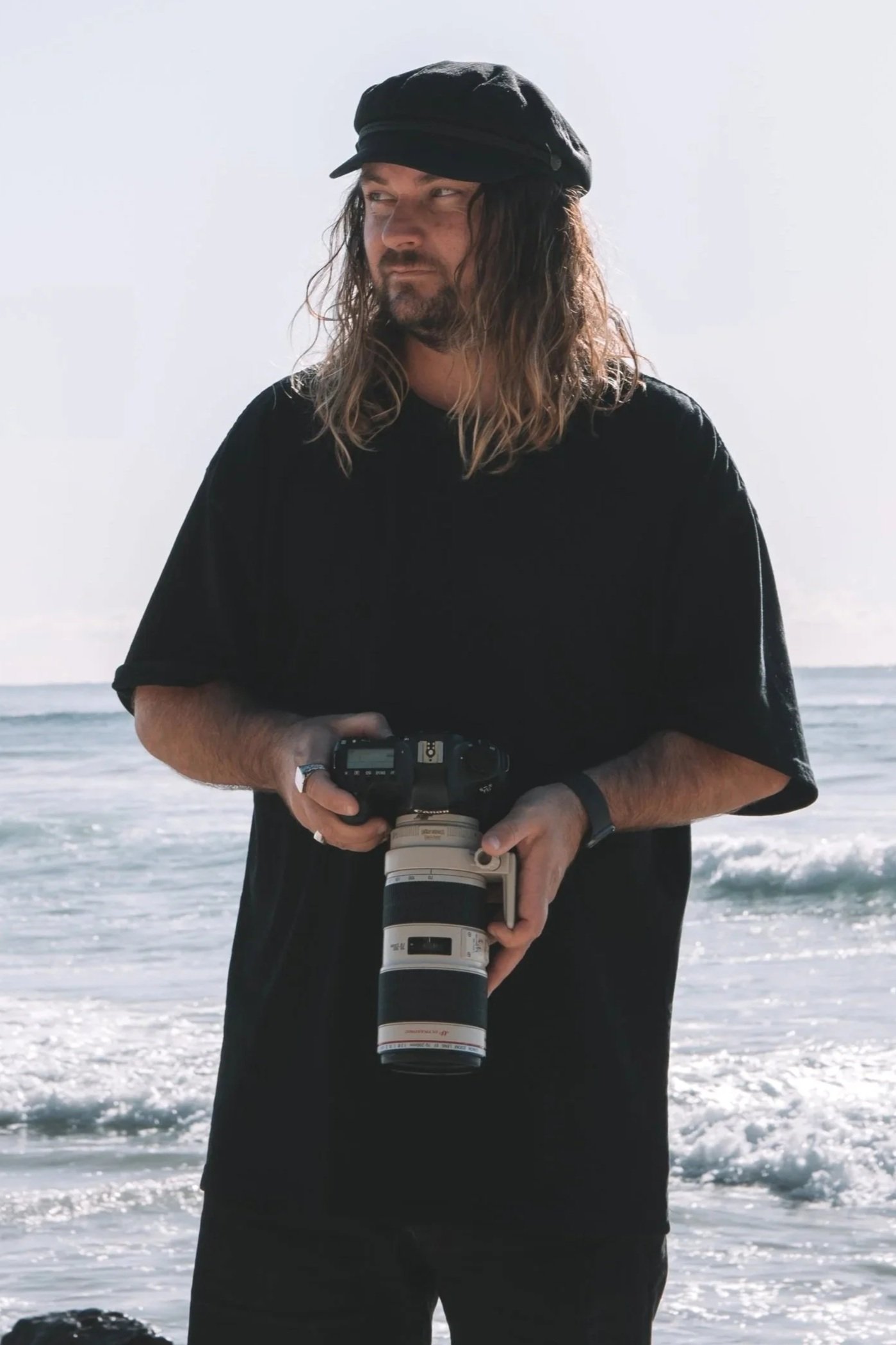 A man with long hair and a beard wearing a black cap and black shirt standing on a beach with ocean waves in the background, holding a camera with a large telephoto lens.