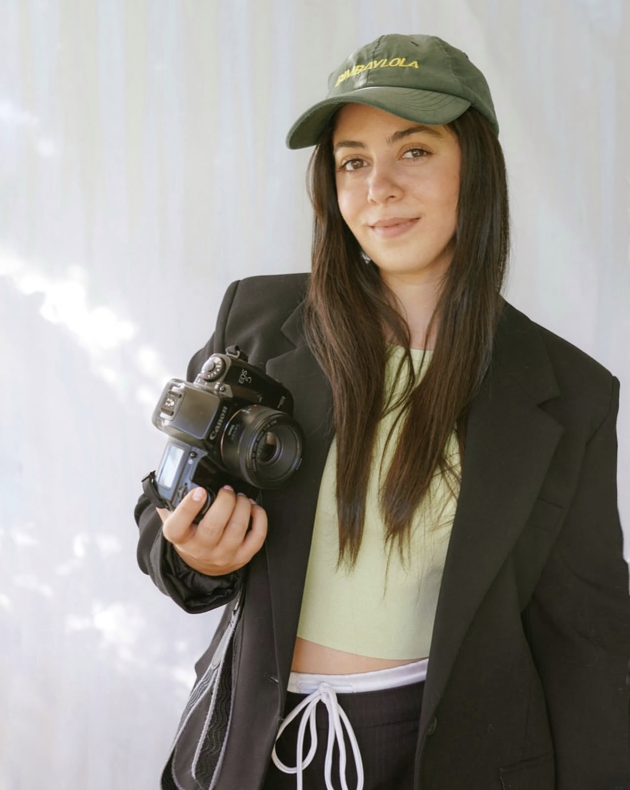 A woman with long brown hair wearing a green cap, black blazer, yellow T-shirt, and black drawstring pants holding a Canon camera.