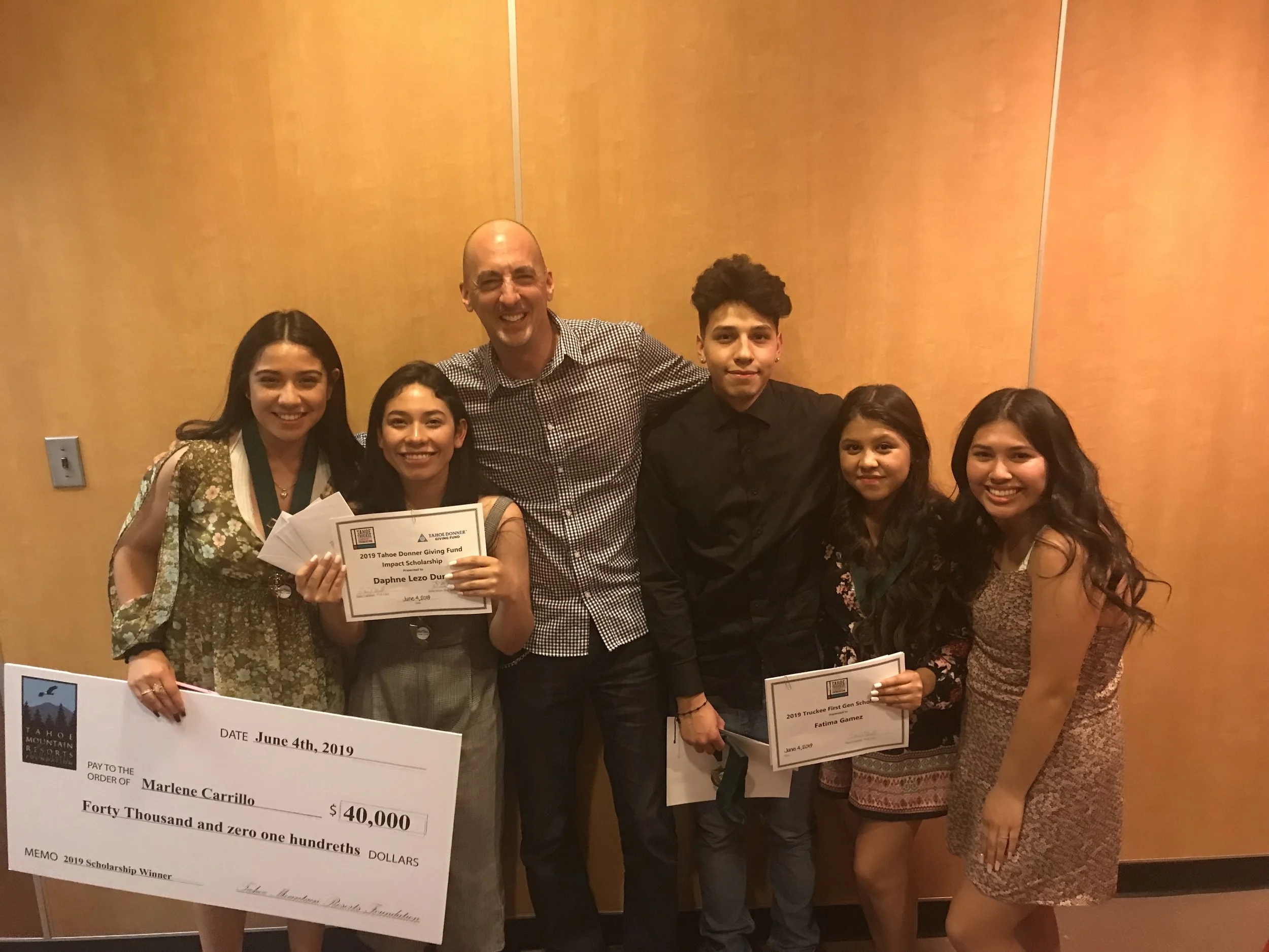Group of six people, four young women and two young men, standing together in front of a beige wall. The women are holding a large check for $40,000 made out to Marlene Carrillo from the Tahoe Donner Giving Fund, and two of the women are holding smal