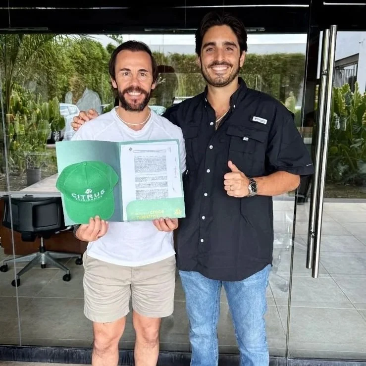 Dos hombres, uno con barba y otro sin, sonriendo y posando frente a una ventana. El hombre con barba sostiene un papel y un gorro verde con la palabra 'CITRUS'.
