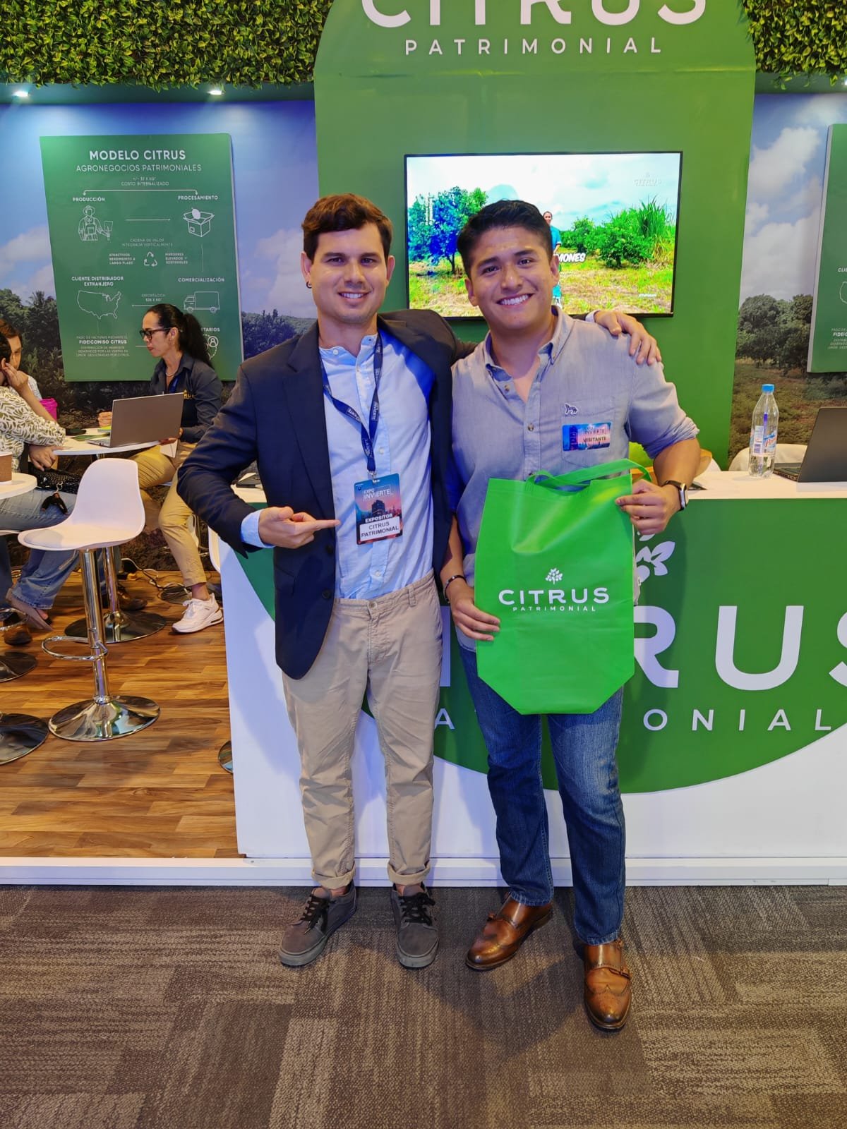 Dos hombres con sonrisas en un evento, uno con traje y el otro con camisa de mezclilla, sosteniendo una bolsa verde con el logo 'Citrus Patrimonial'.