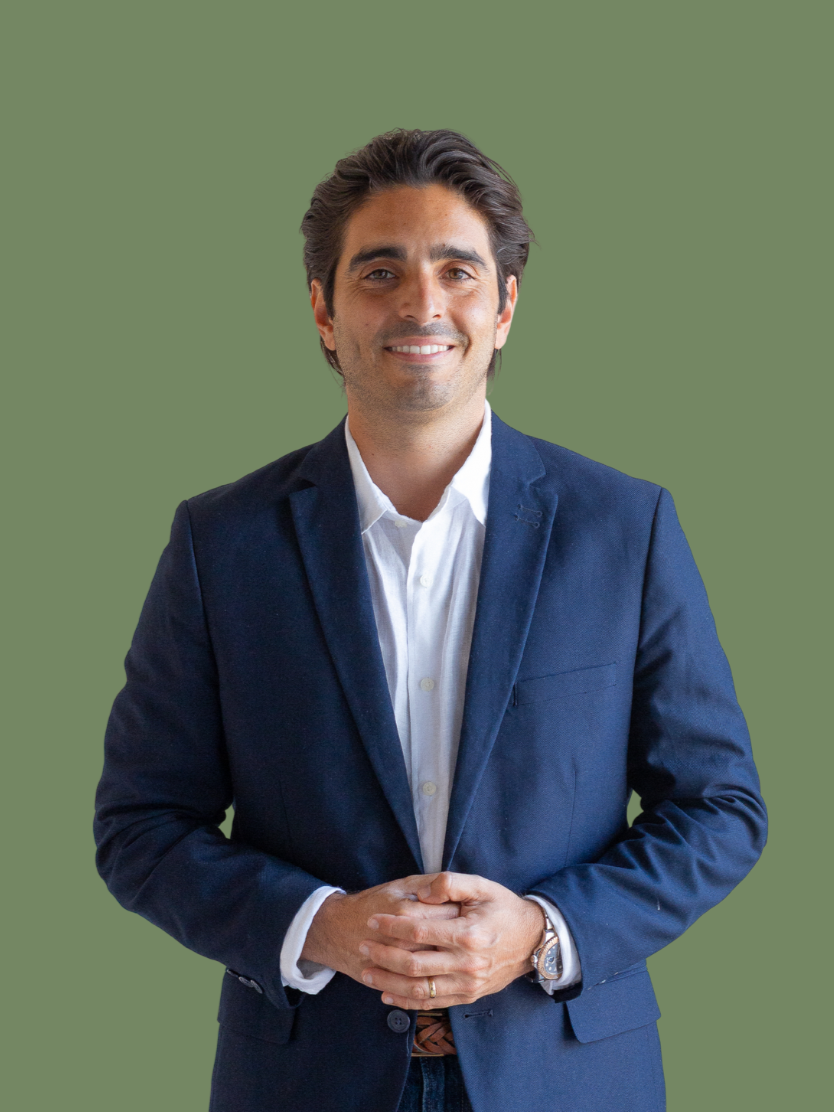 Hombre sonriente con barba, cabello oscuro y peinado hacia atrás, vistiendo un saco azul oscuro y una camisa blanca, posando con las manos juntas contra un fondo verde.