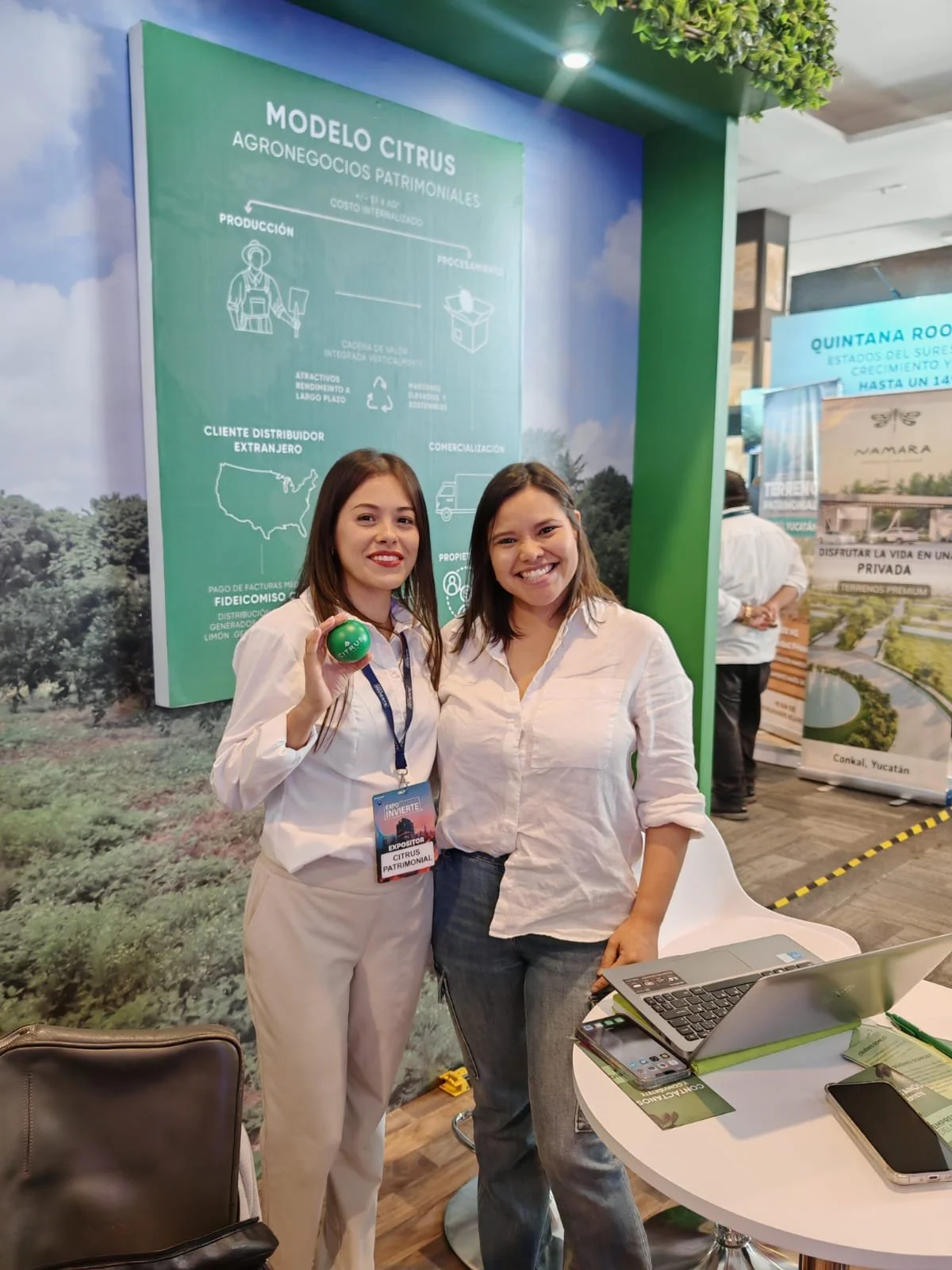 Dos mujeres sonriendo en un stand de una exposición, una de ellas sostiene una pelota verde. Hay una mesa con laptop y teléfonos, y un cartel en el fondo que dice 'Modelo Citrus Agronegocios Patrimoniales'.